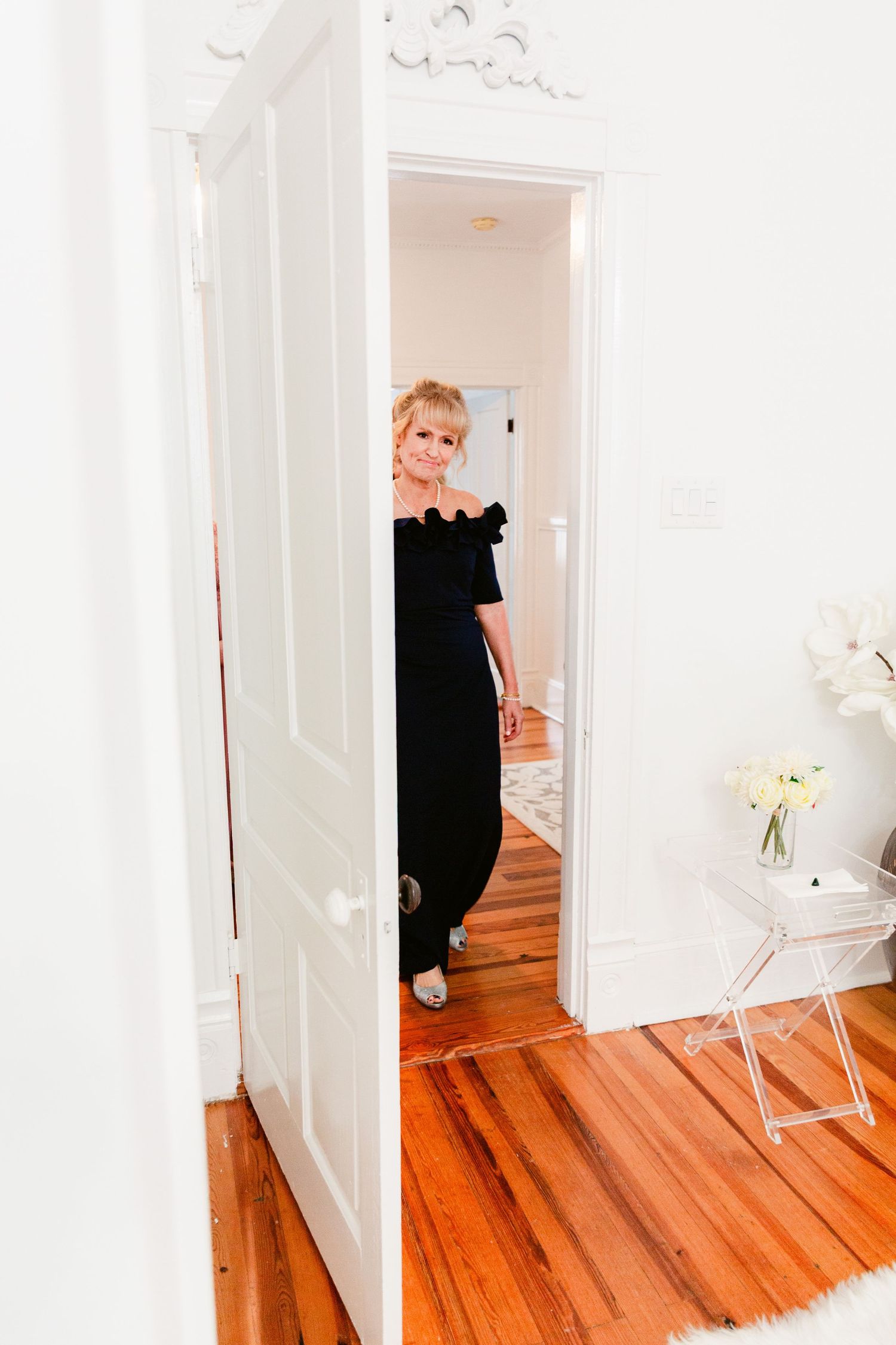 Woman in elegant black off-shoulder dress stands in doorway of bright white room with polished hardwood floors.