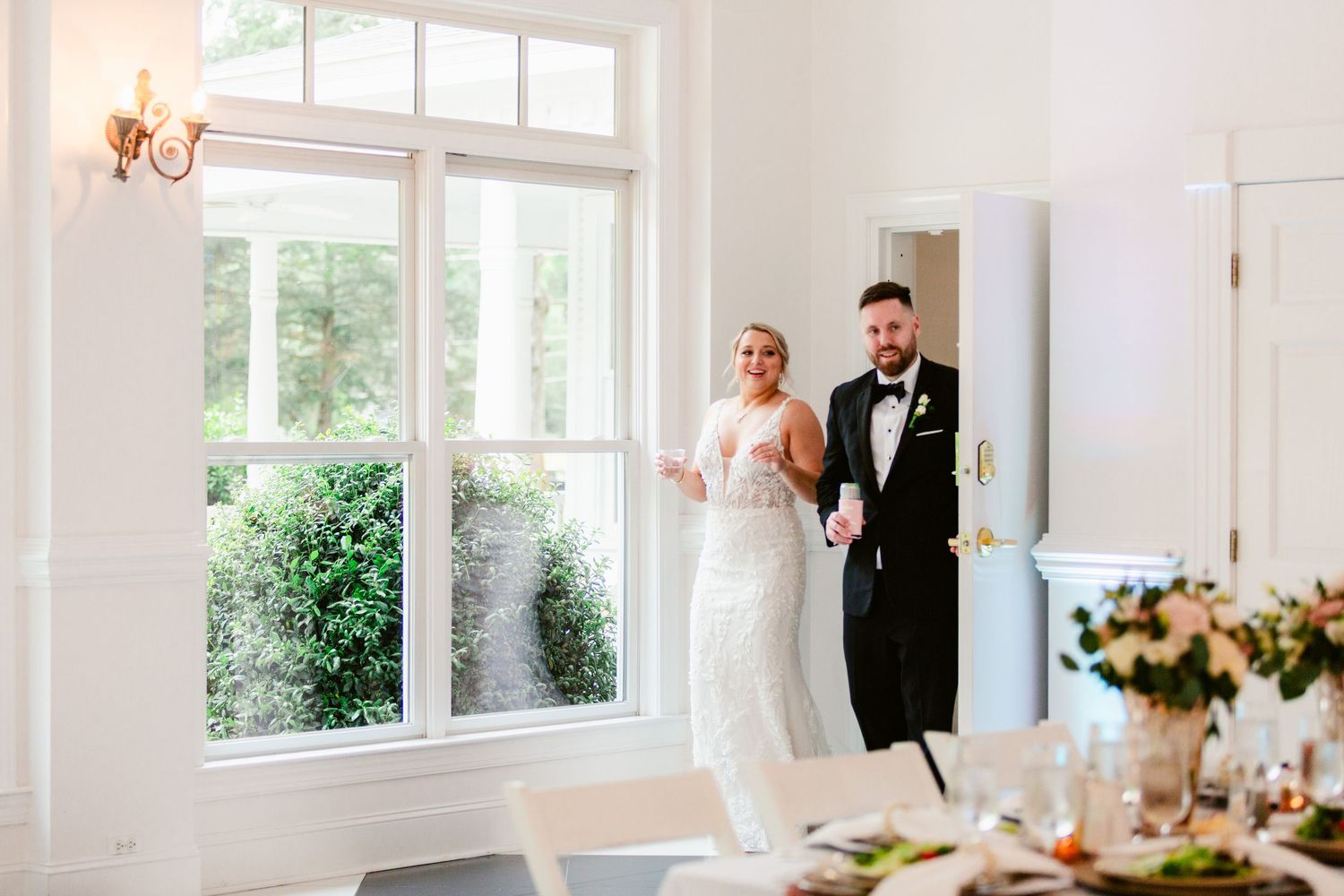 Elegant white wedding reception venue with large windows, floral centerpieces, and natural light streaming in.