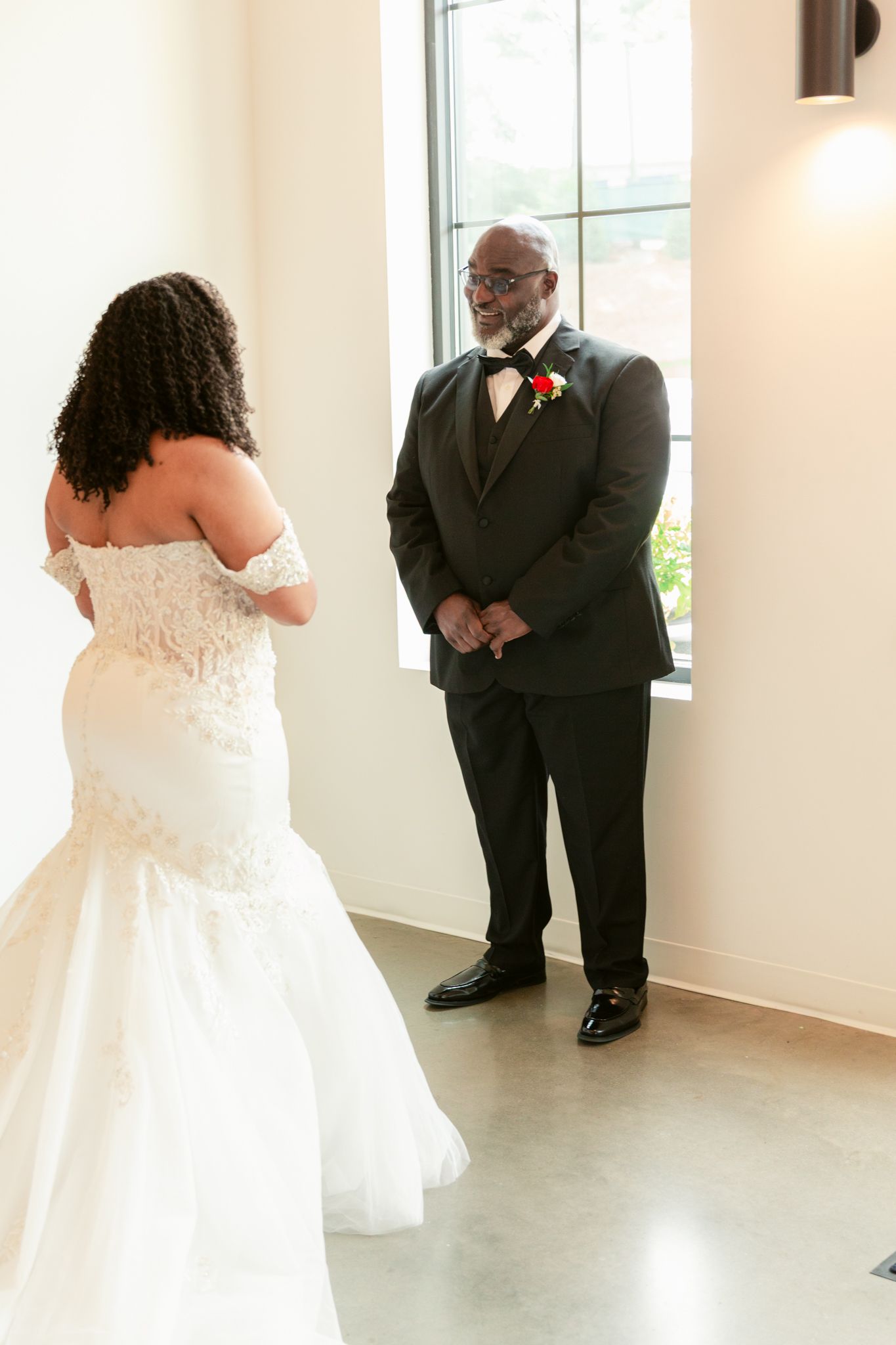 Emotional first look moment between a bride in a strapless lace gown and someone in a dark suit at an indoor venue.