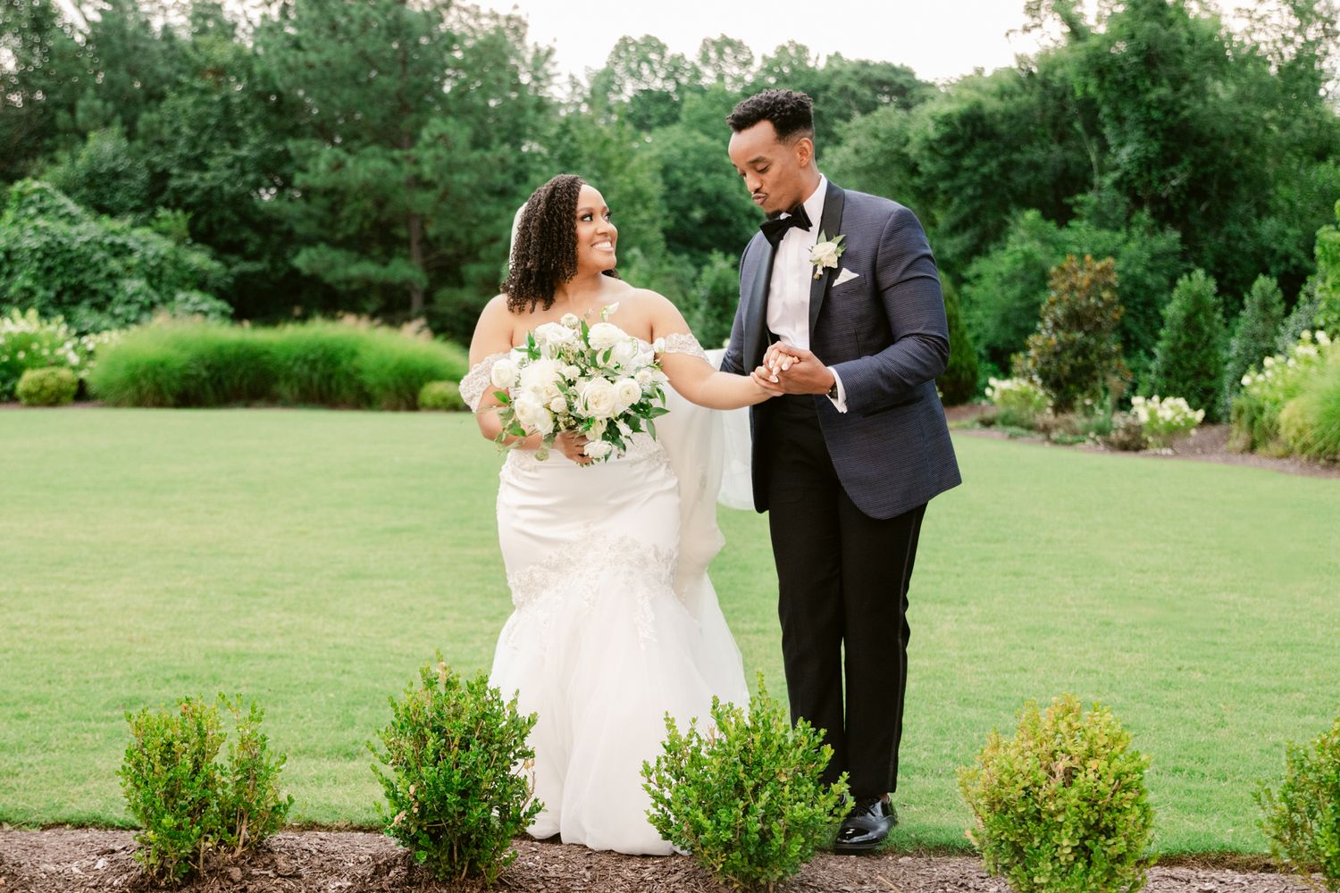 A couple in wedding attire shares a romantic moment in a lush green garden setting.