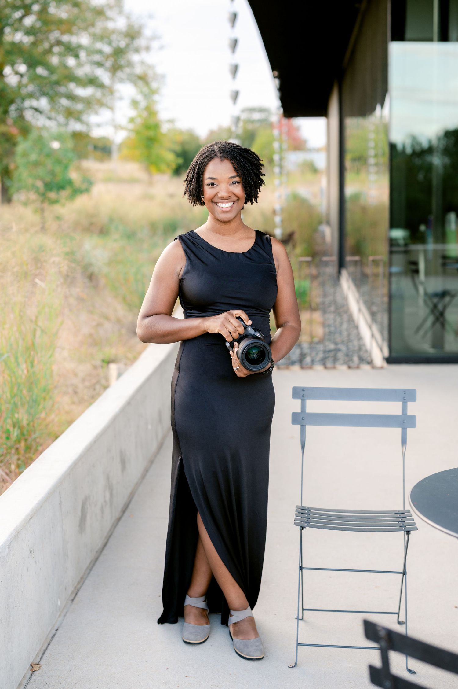 A photographer in a black dress poses with a camera on an outdoor patio with blue chairs.