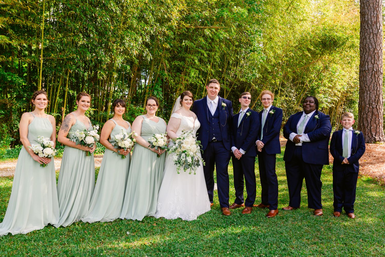 Wedding party in formal attire stands in line on green lawn with trees in background during golden hour.