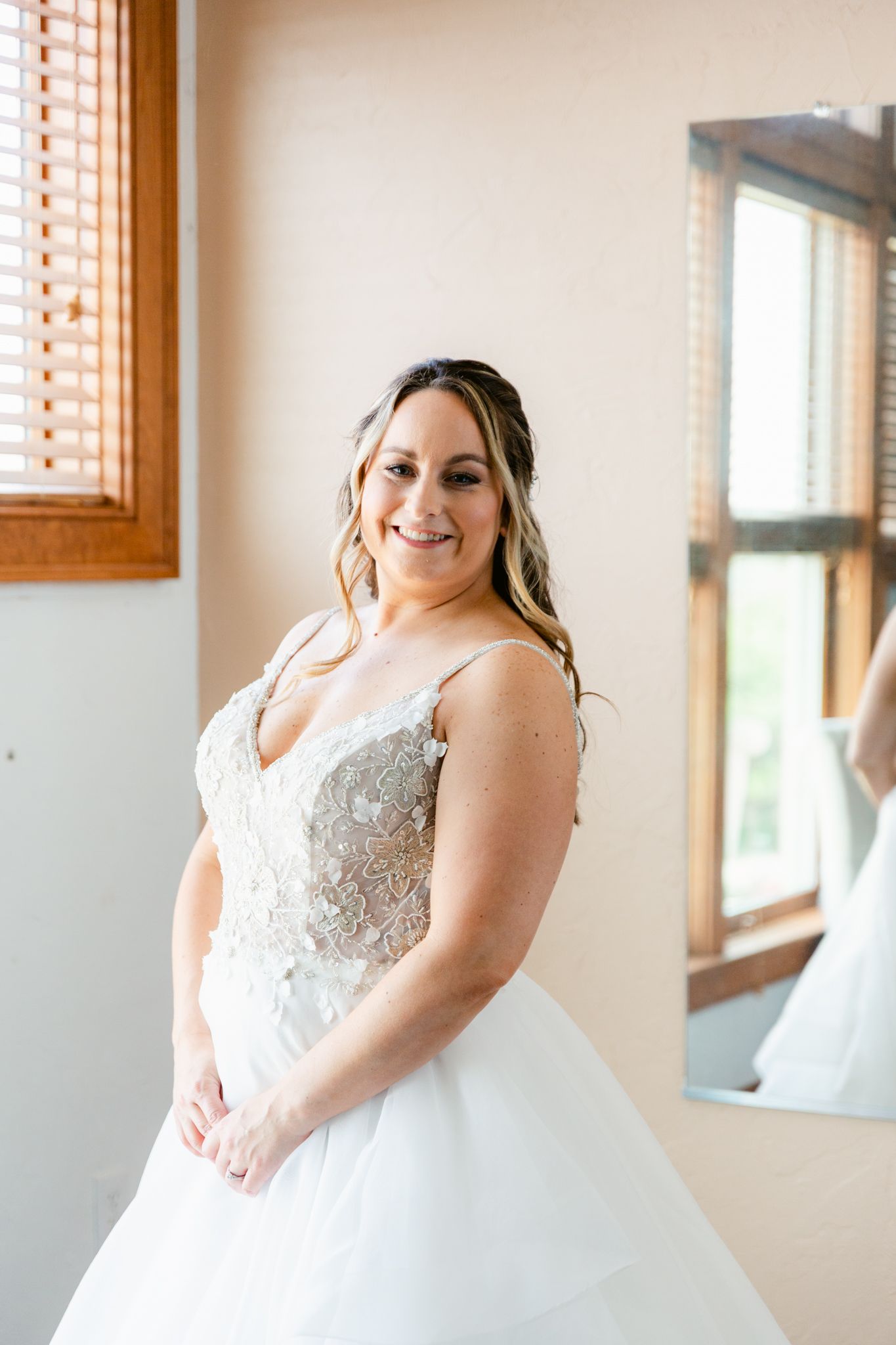 Bride in elegant white lace wedding dress smiles while standing near window in natural light.