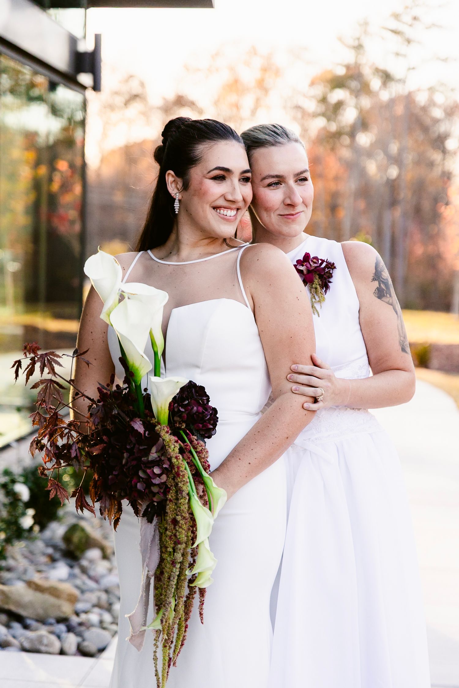 Two people in white dresses embrace while holding deep burgundy and green floral bouquets at an outdoor wedding.