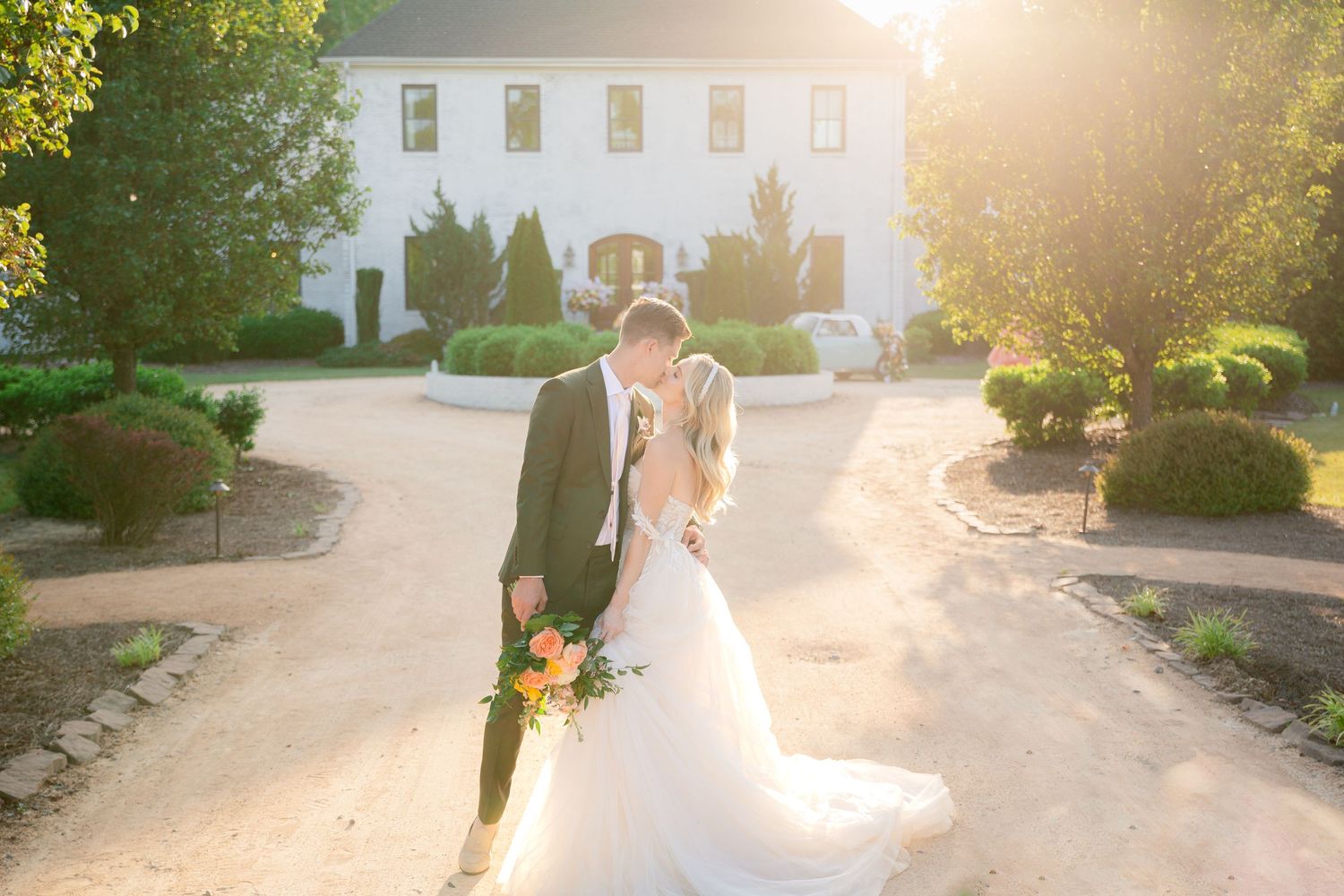 Wedding couple walks down a sunlit path at sunset with a white colonial building in the background.