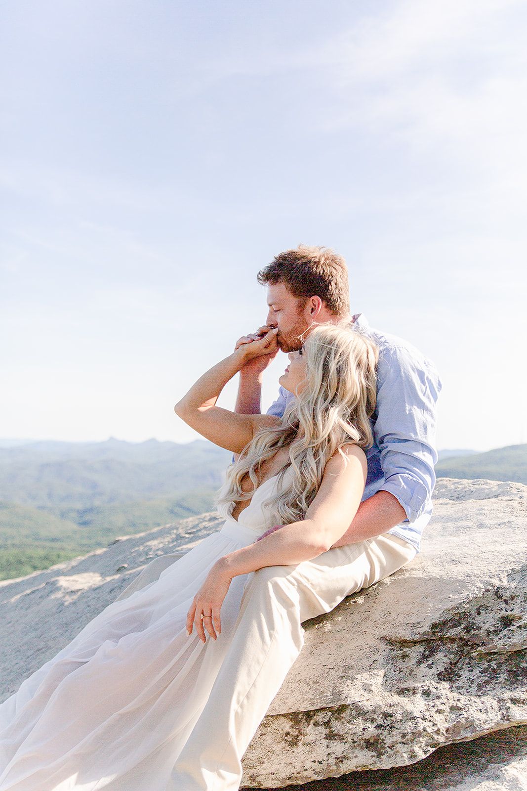 Couple in casual attire pose romantically on rocky mountain overlook with scenic vista.