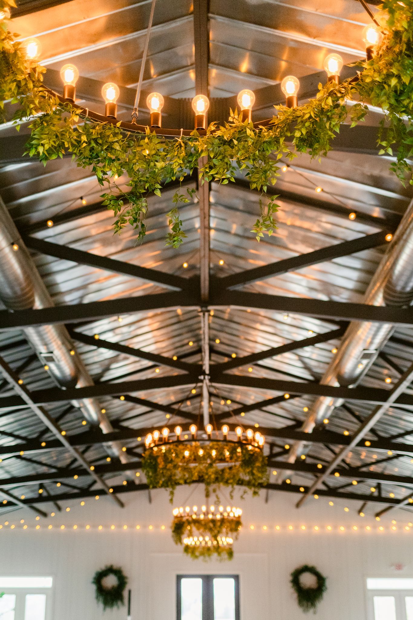 Elegant wedding venue ceiling with exposed beams, string lights, chandeliers and greenery garlands creating romantic ambiance.