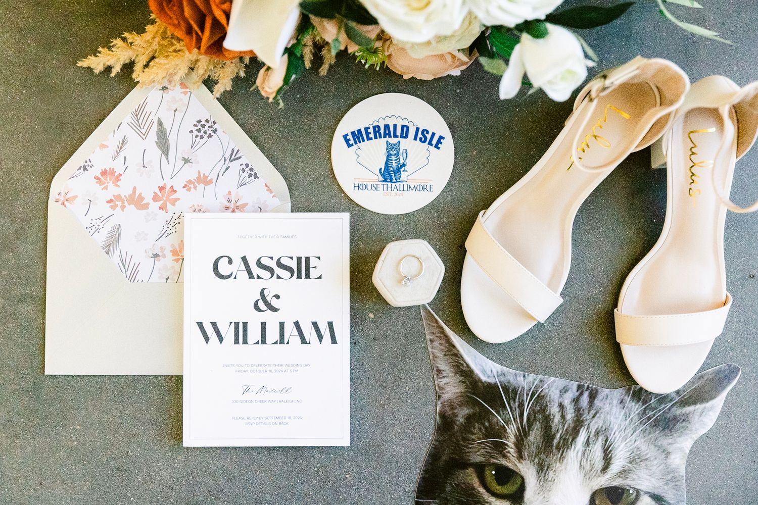 Wedding details flatlay with ivory heels, invitation card, ring box, and gray cat peeking into frame.