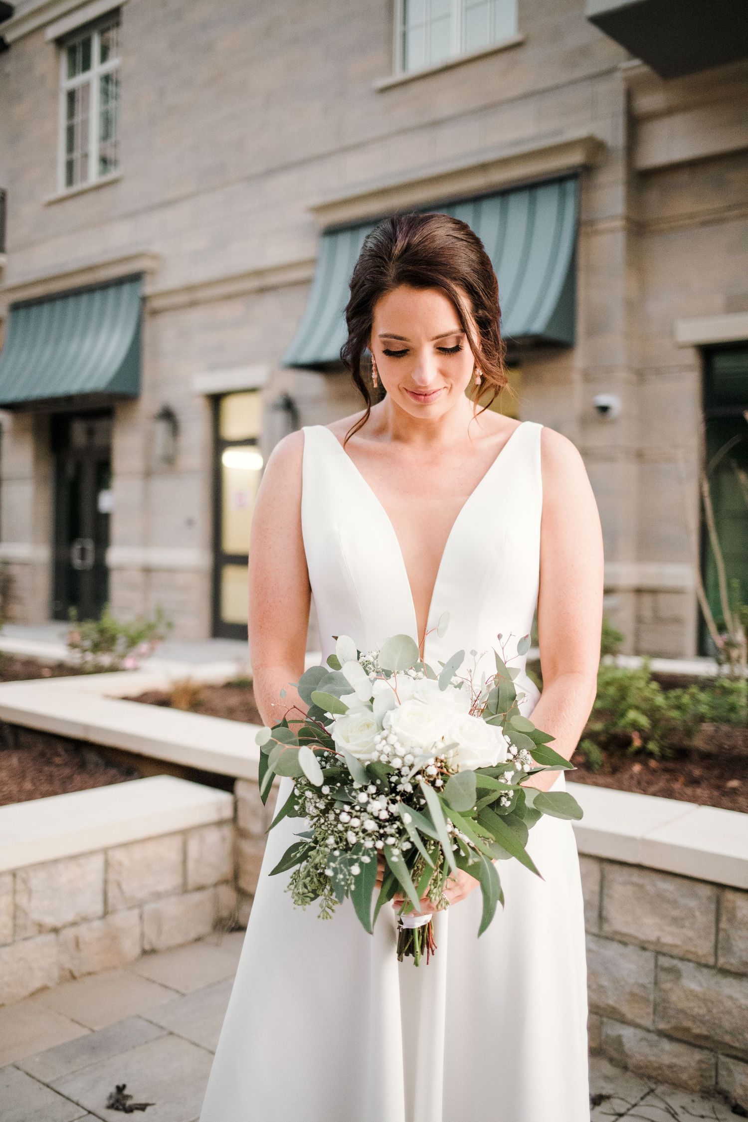 Elegant bride in v-neck wedding dress holds white and green bouquet against stone building.