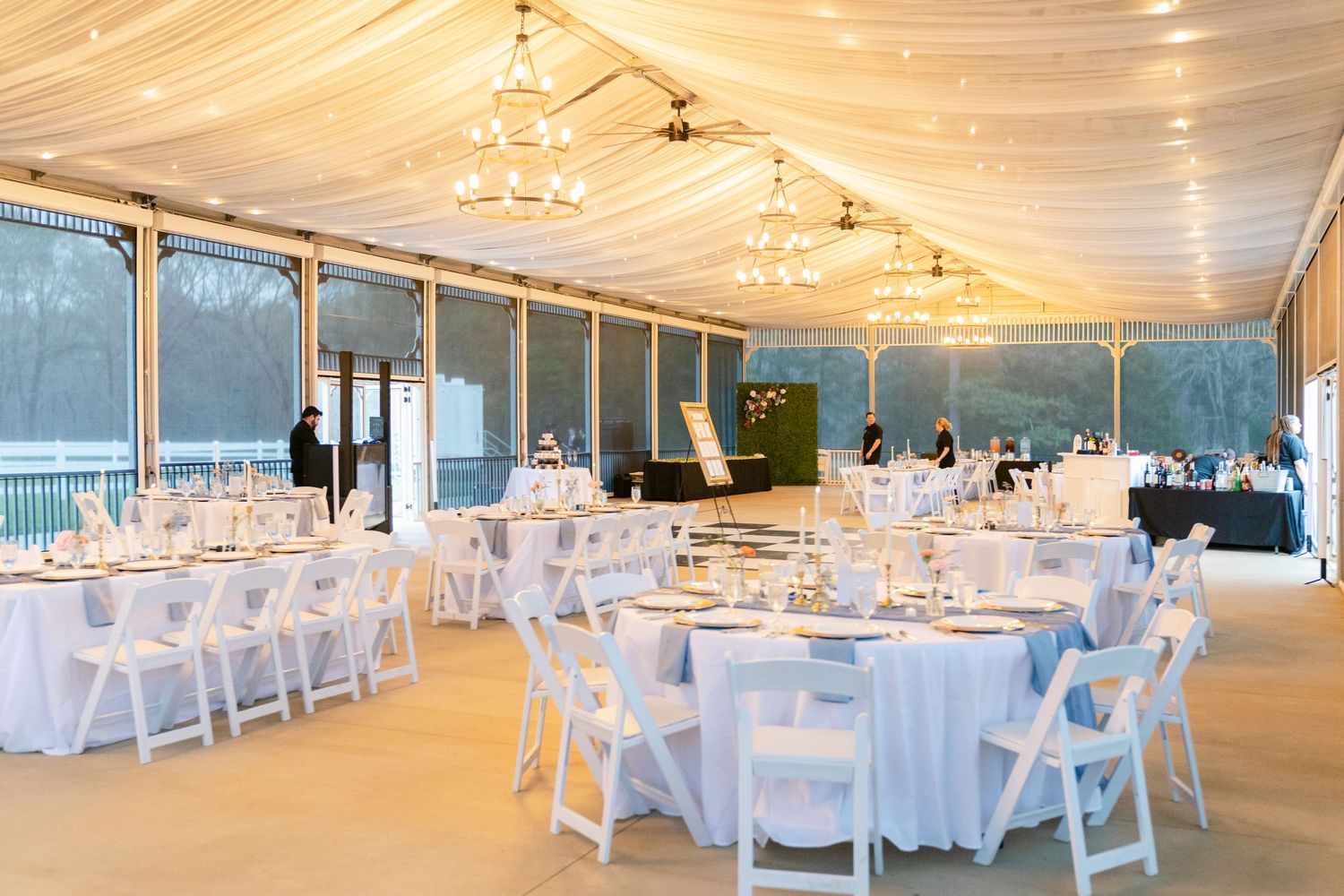 White tent reception venue setup with round tables, white folding chairs and string lights draped across ceiling.