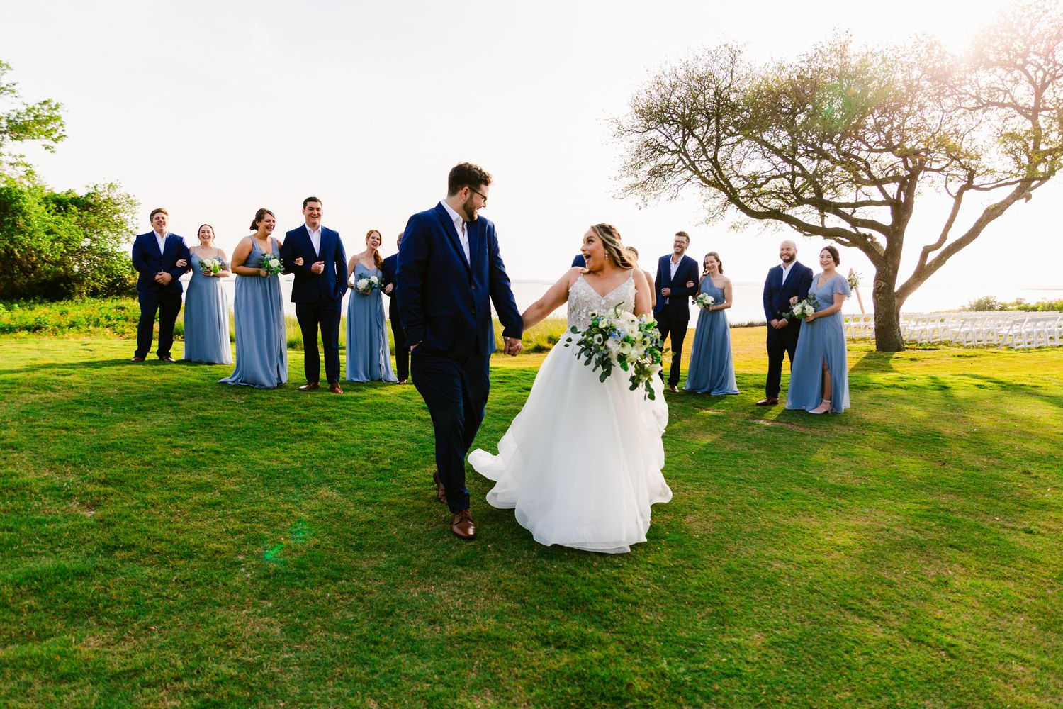 Wedding party members gather on a grassy hill overlooking a scenic waterfront view during an outdoor celebration.