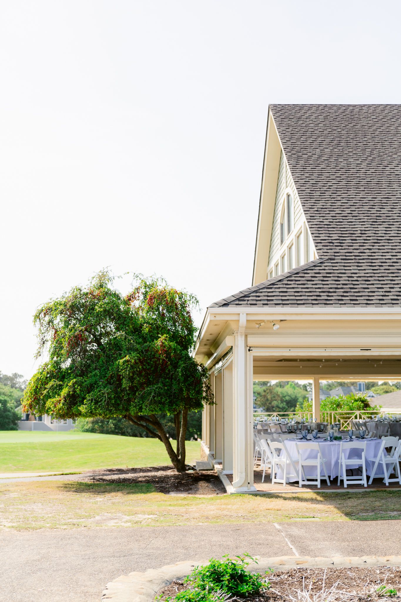 White wedding chairs arranged outdoors near a rustic chapel with a lush green tree providing natural shade.