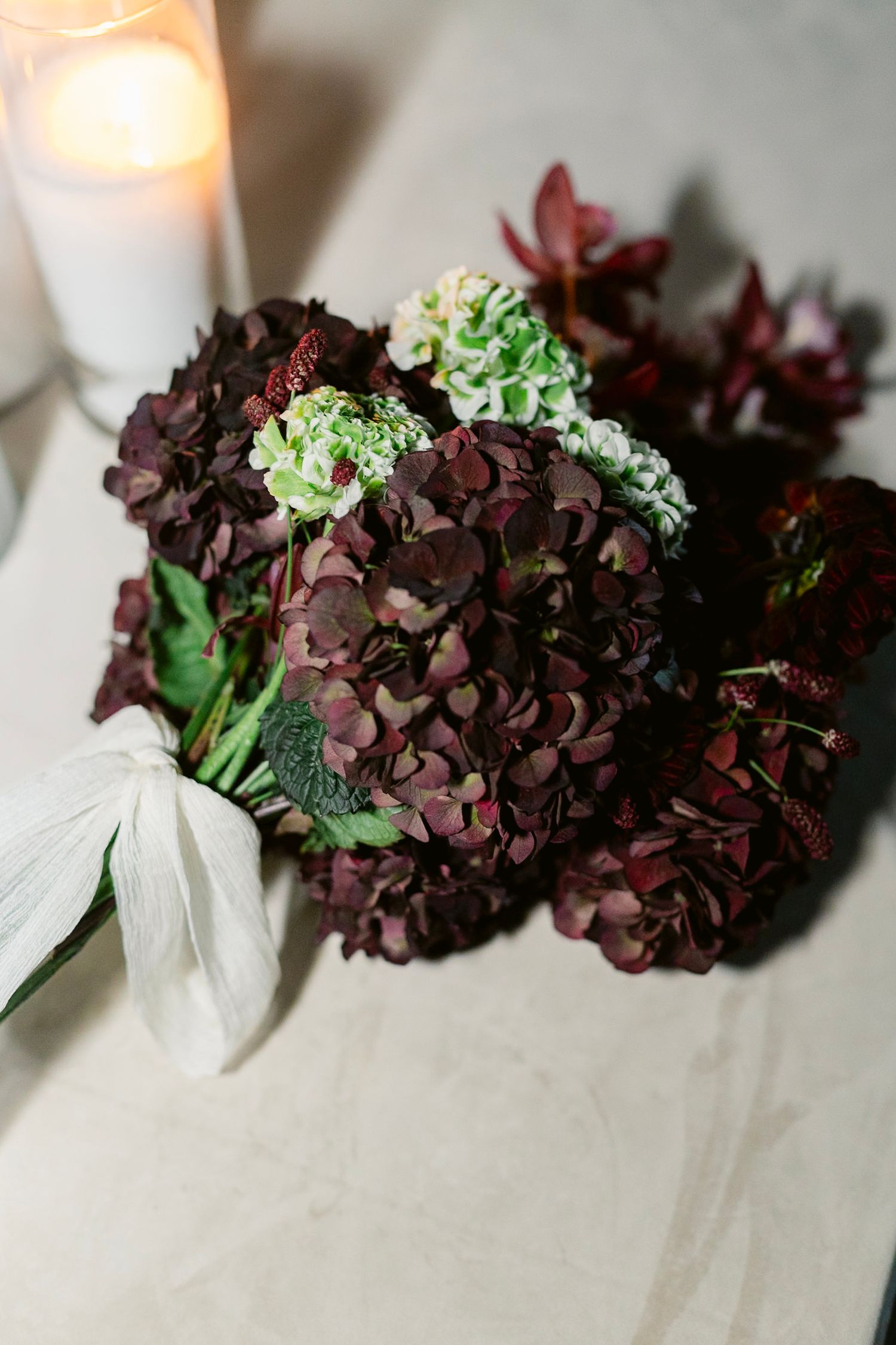 Deep burgundy hydrangea blooms with light green accents and white ribbon detail in moody lighting with a candle.