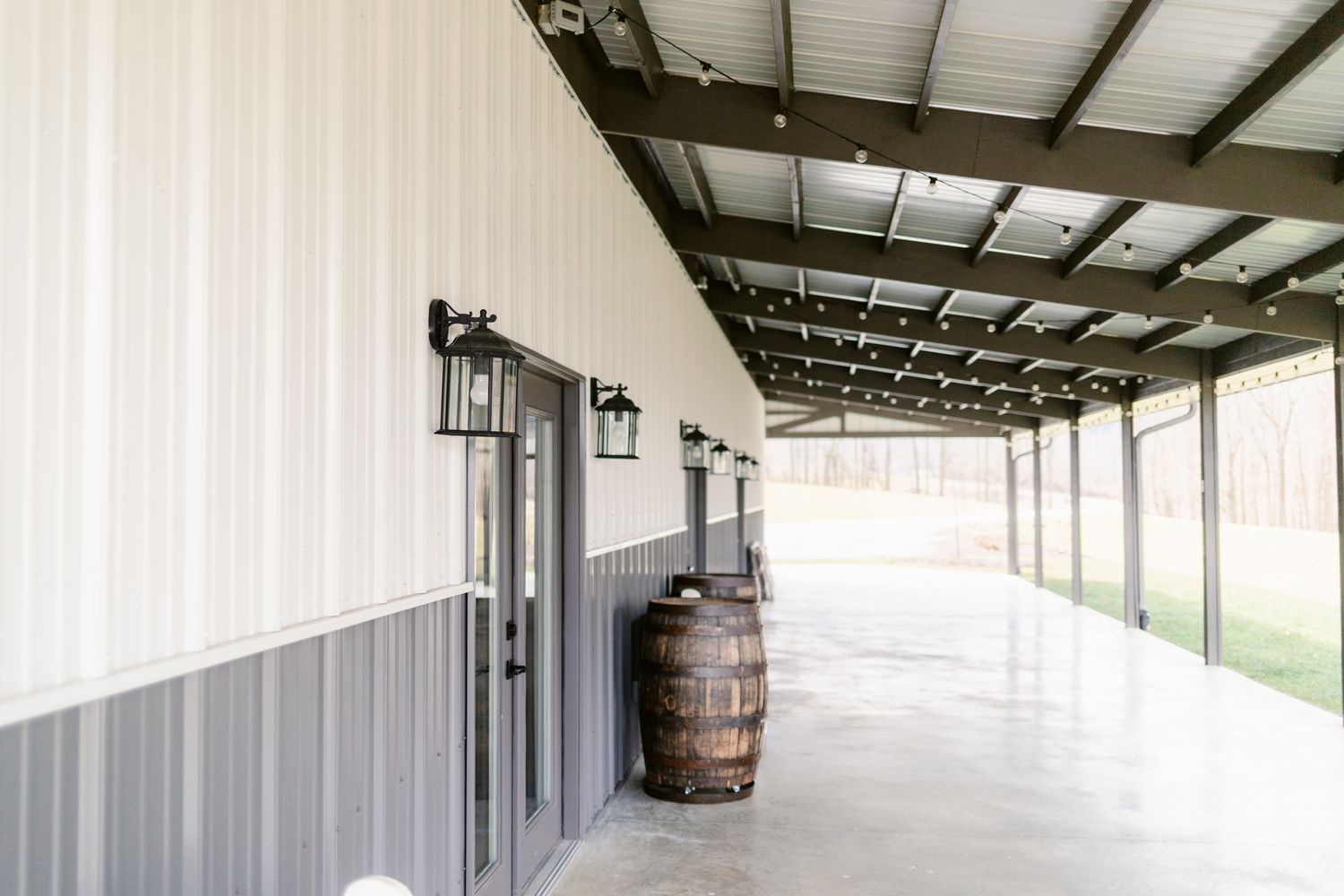 Long covered walkway with white walls and wooden barrels along a rustic building exterior.