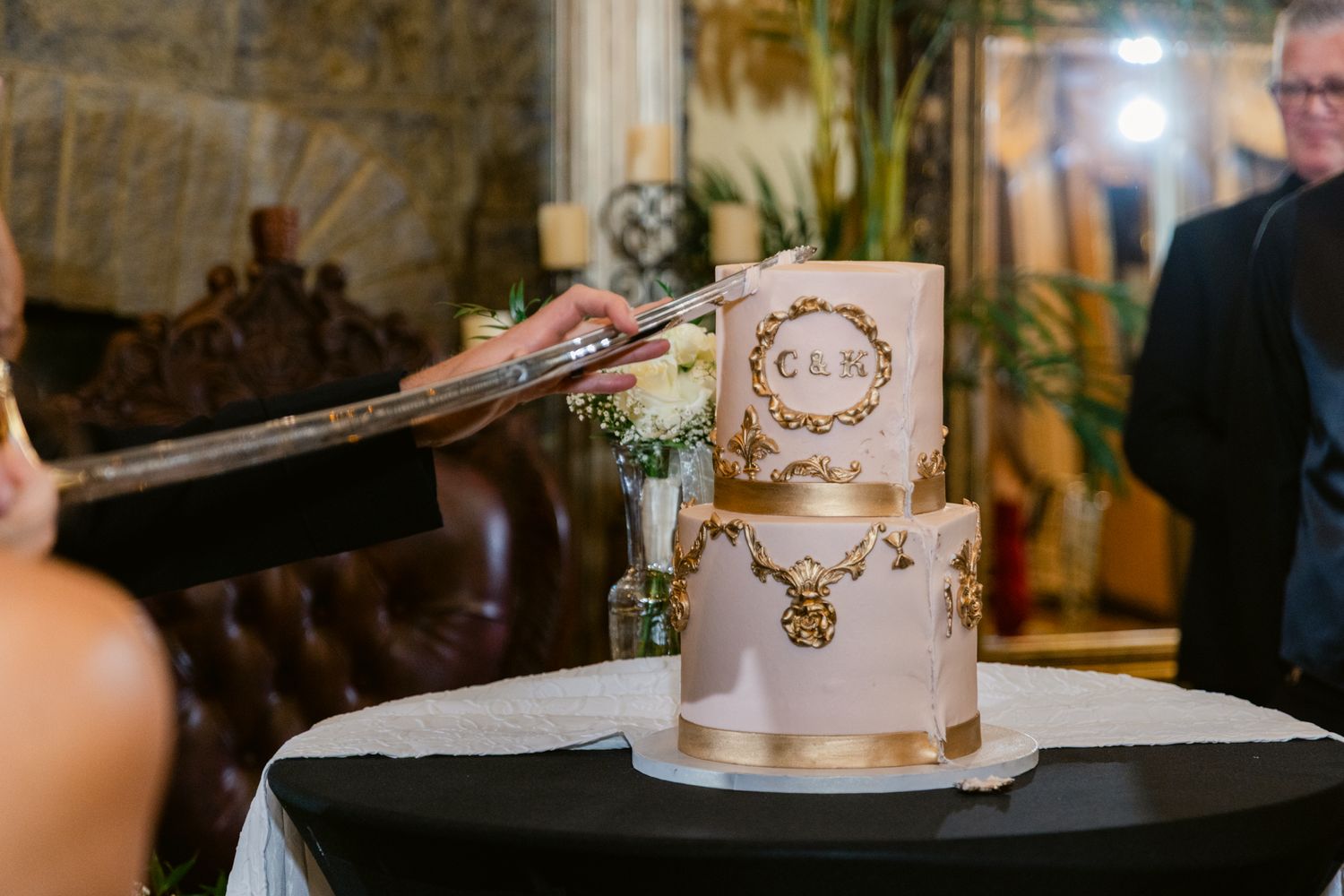 An elegant two-tier white and gold wedding cake with a monogram design sits on a black tablecloth at a reception.