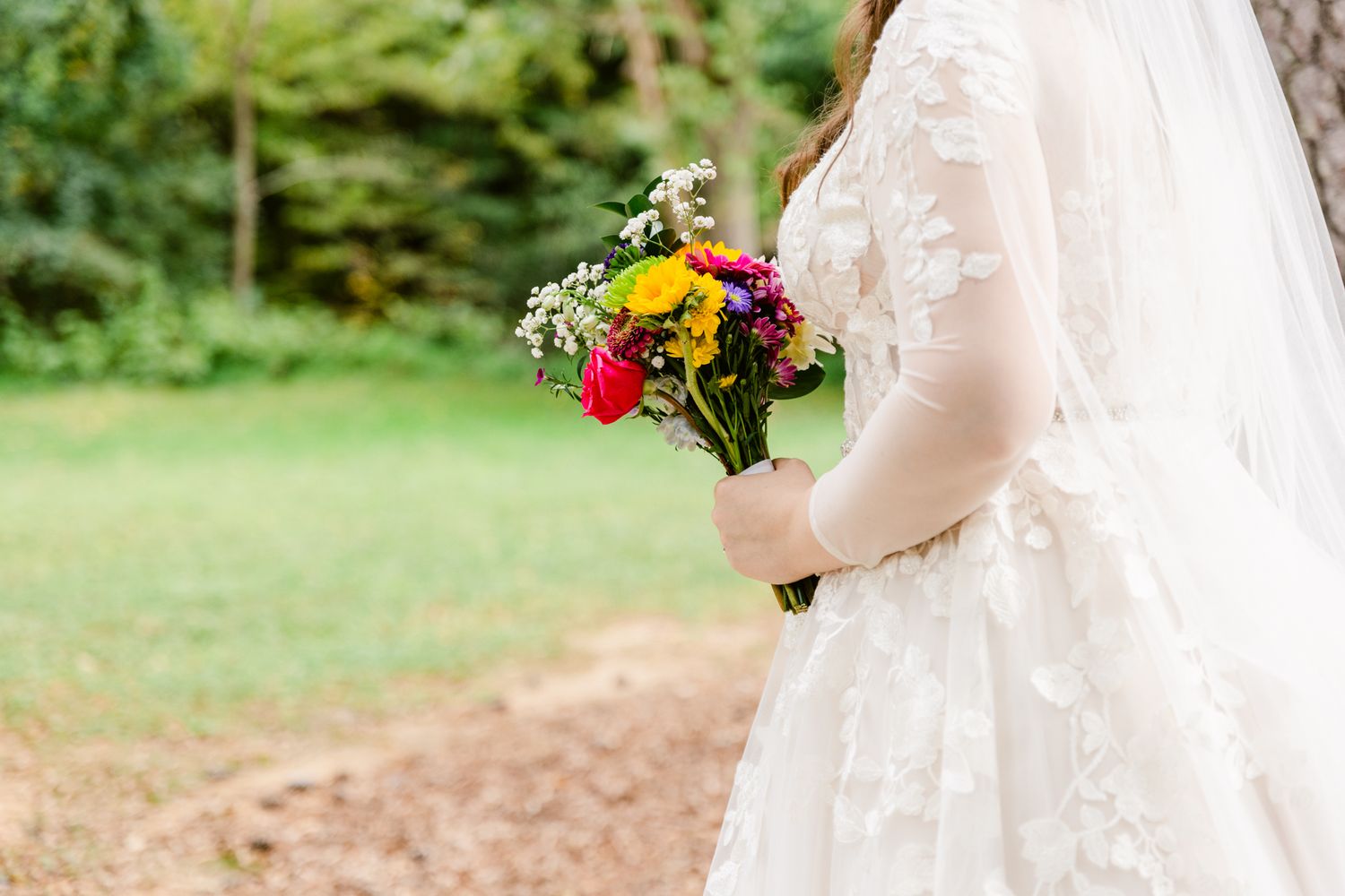 A bride holds a vibrant bouquet of yellow and pink flowers against a white lace wedding dress in an outdoor setting.