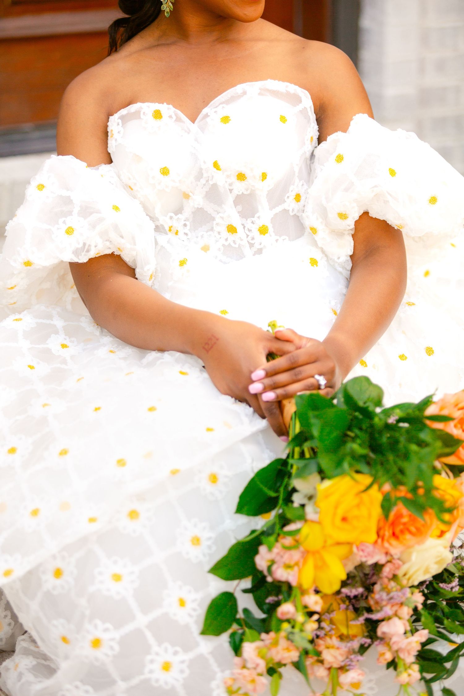 A bride in a white daisy-patterned wedding dress holds a vibrant yellow and pink floral bouquet.