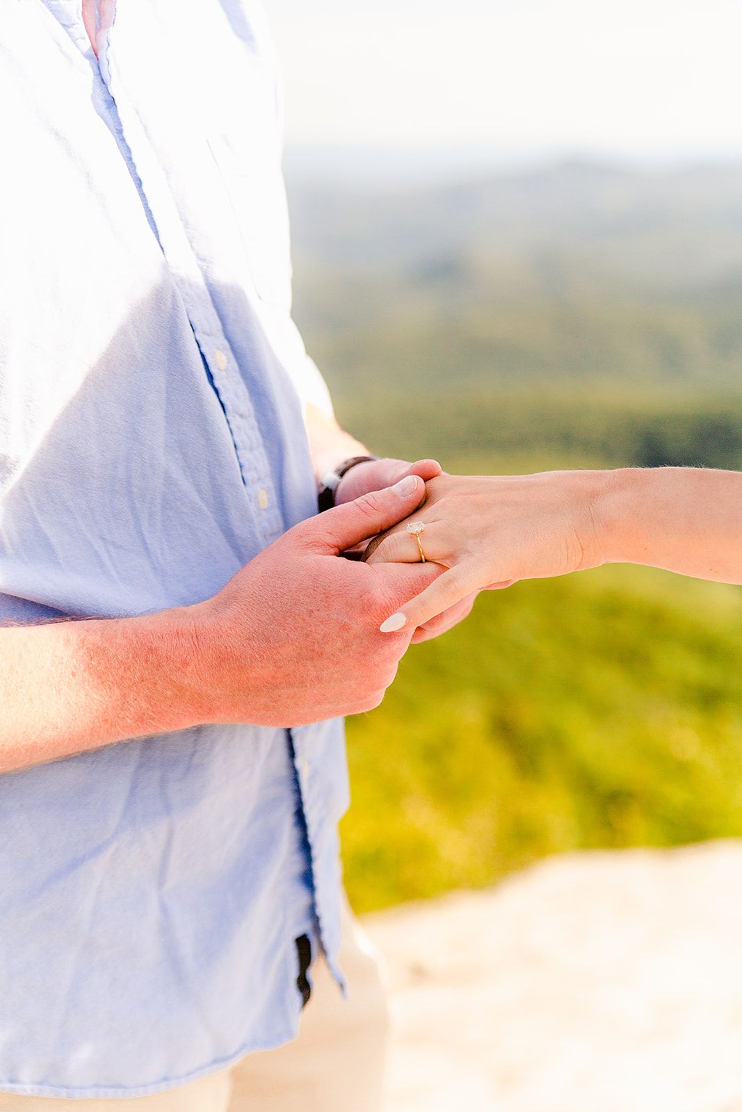Close up of hands clasped together wearing a wedding ring against a blurred outdoor landscape background.