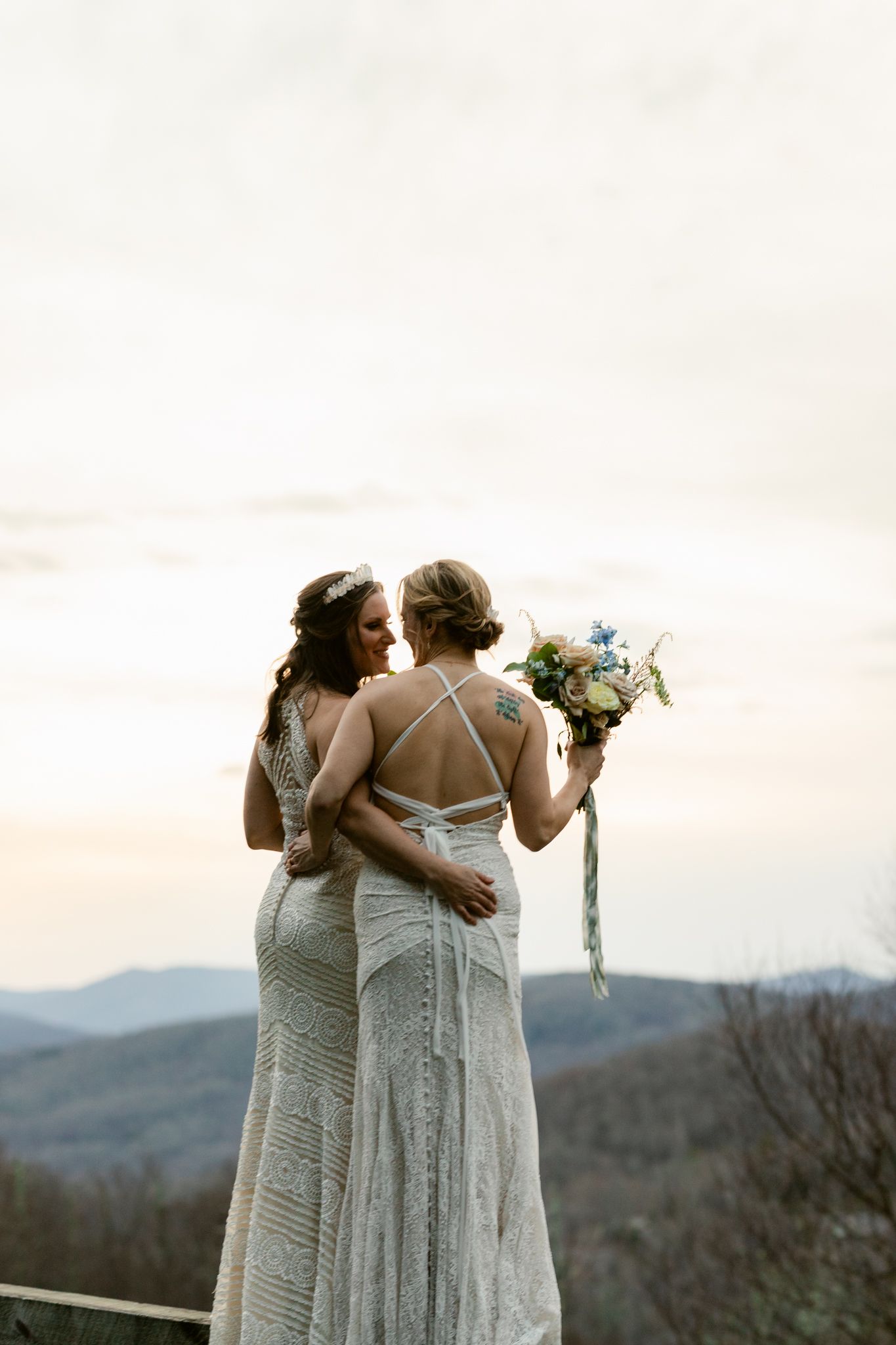 Three people wearing matching white lace dresses pose together against a mountain landscape backdrop.