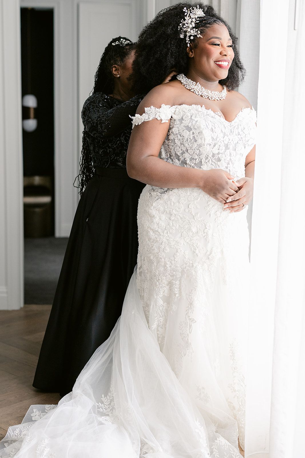 Beautiful bride wearing an off-shoulder lace wedding gown with crystal tiara smiles while getting ready by a window.