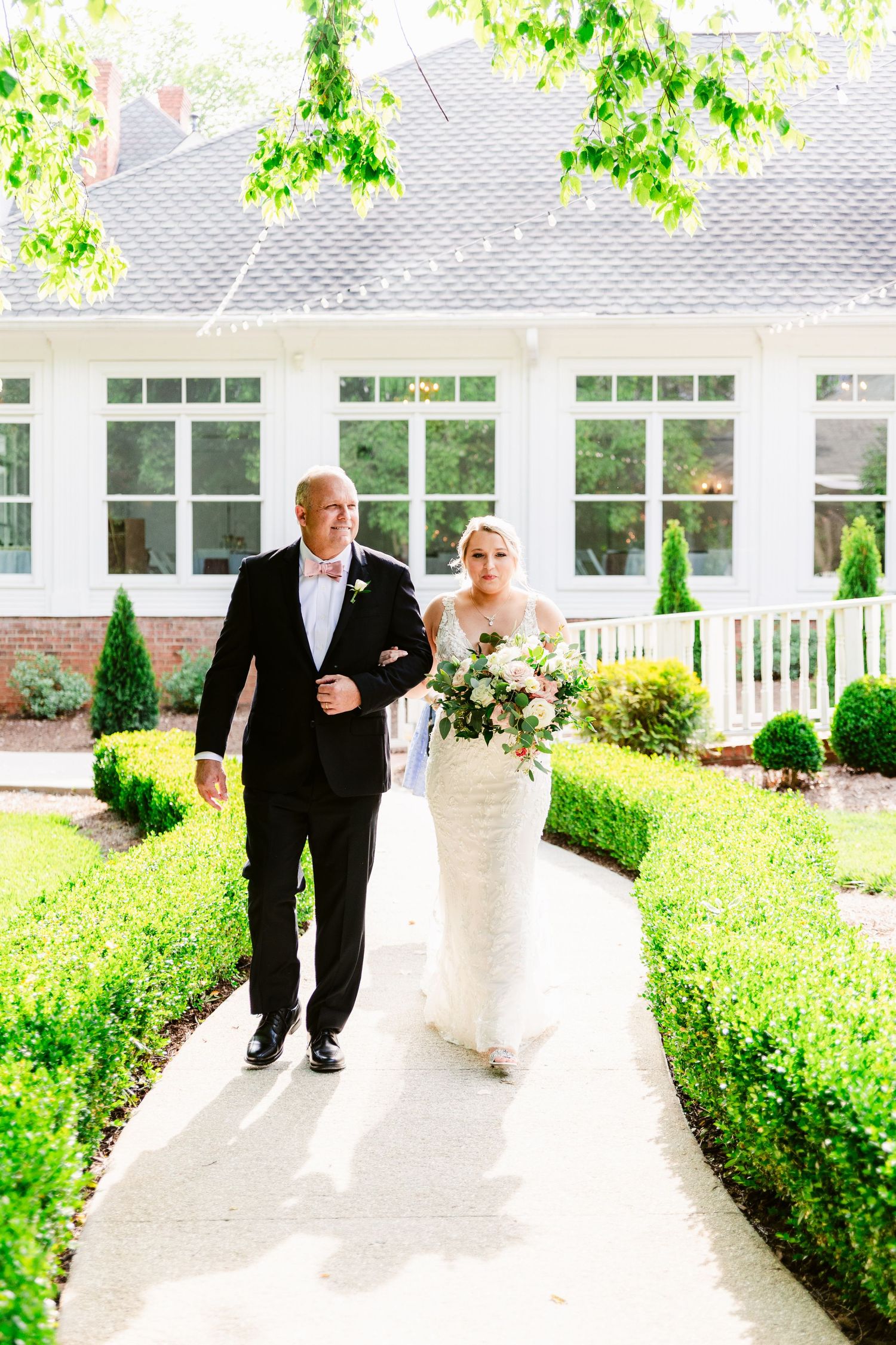 A wedding party walks along a white columned walkway surrounded by lush green landscaping on a sunny day.