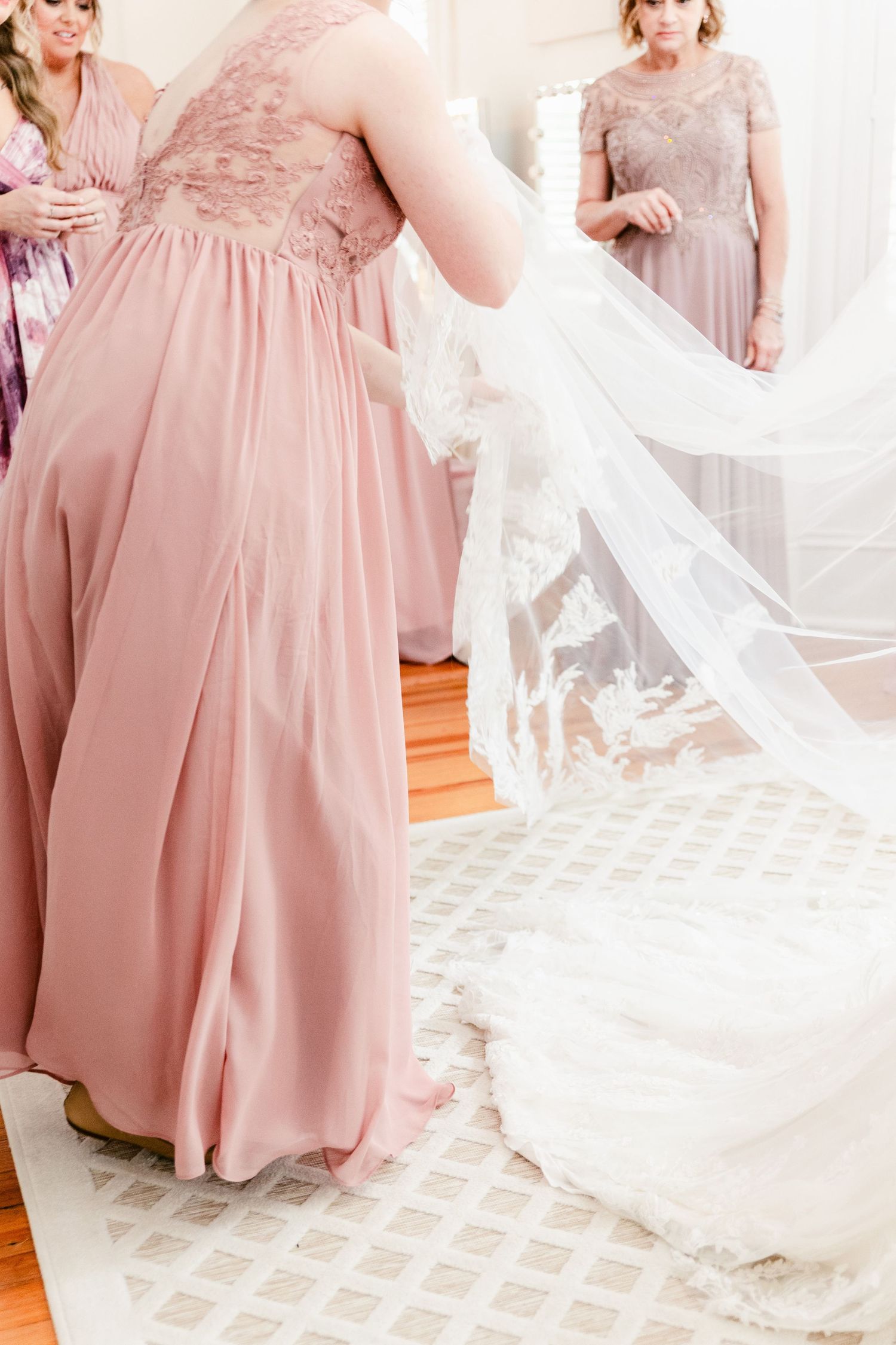 Bridesmaids in blush pink dresses gather around a bride in a flowing white gown with delicate lace details.