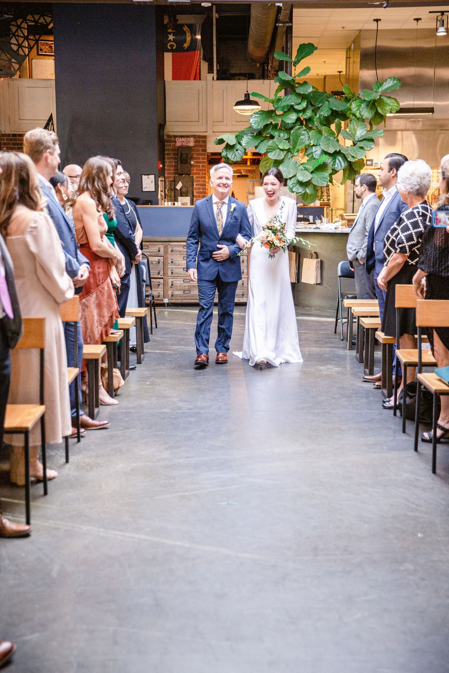 A wedding ceremony takes place in a modern industrial venue with high ceilings and a large potted plant as decor.