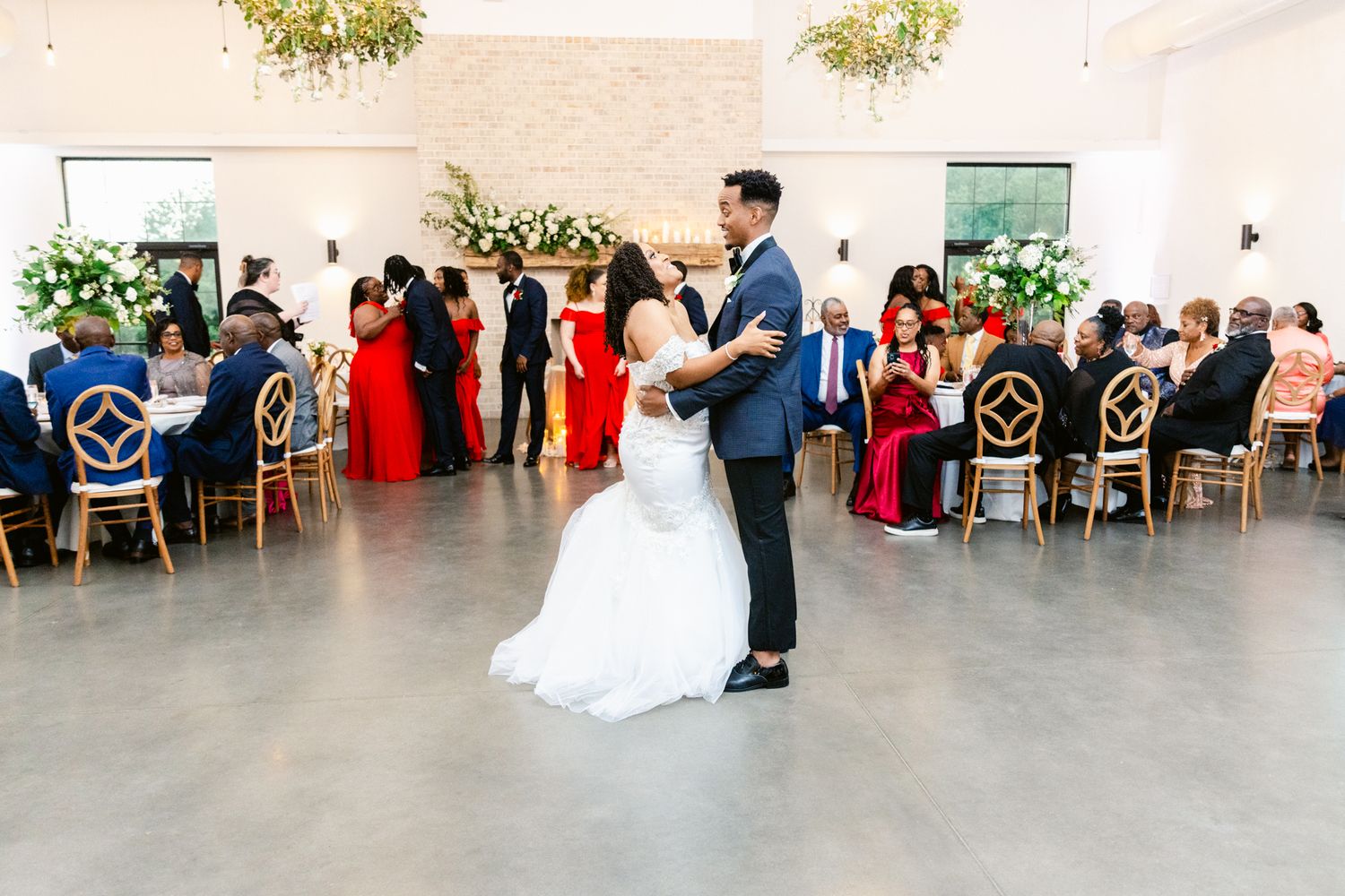 Wedding guests seated at long tables in an elegant white reception venue with natural lighting.