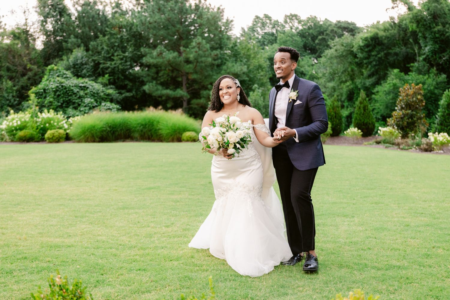 A happy wedding couple walks together on a beautiful garden lawn holding bouquets.