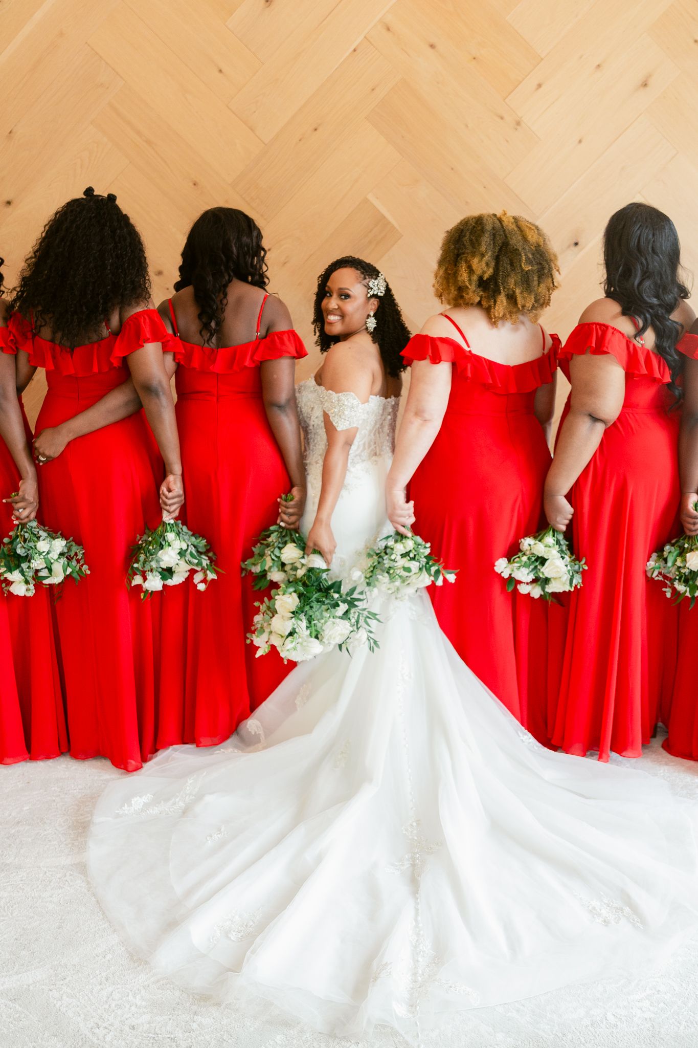 A bride in white gown stands with bridesmaids wearing matching red dresses holding white bouquets against a beige wall.