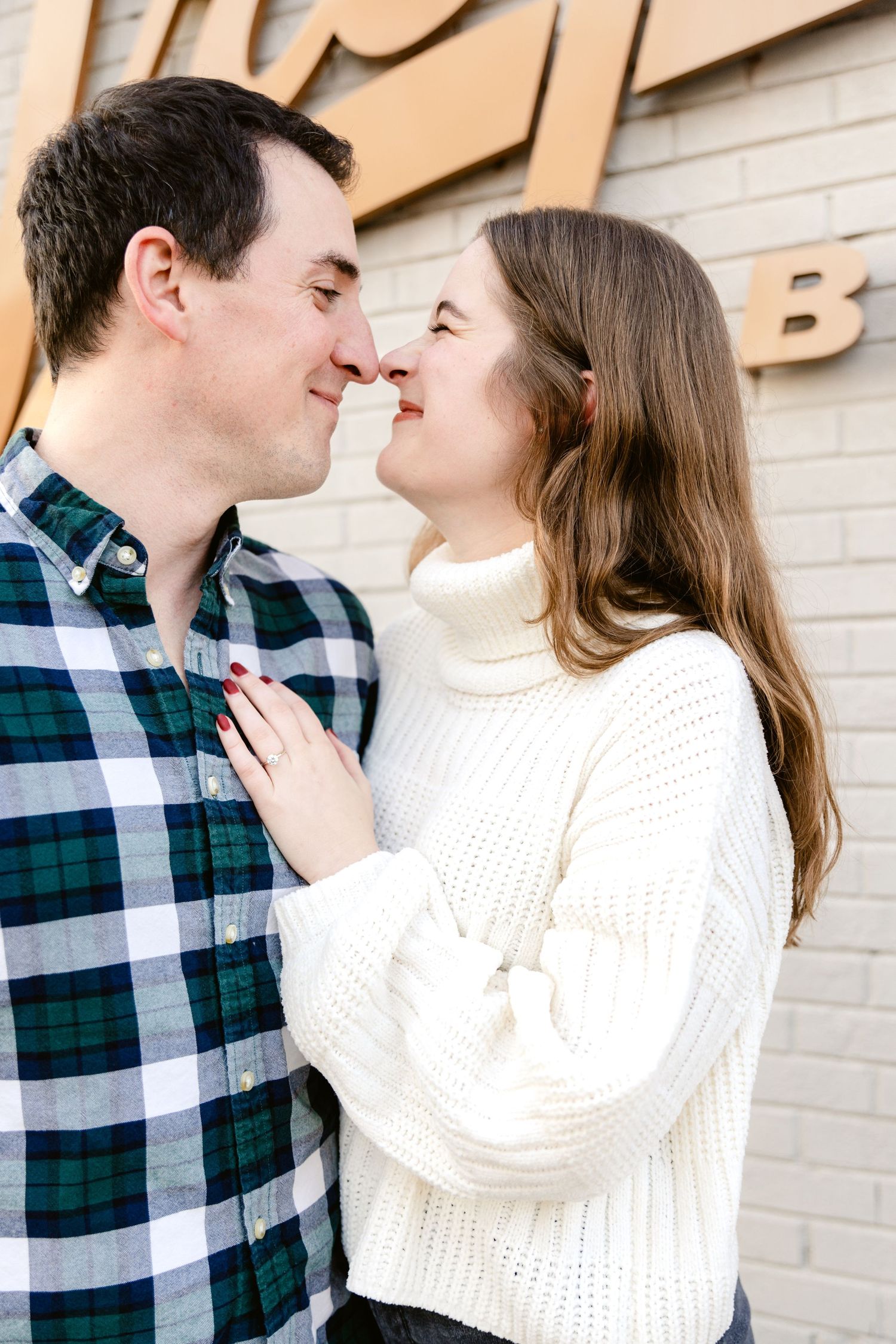 A couple in casual winter attire share an intimate moment against a beige wall background.