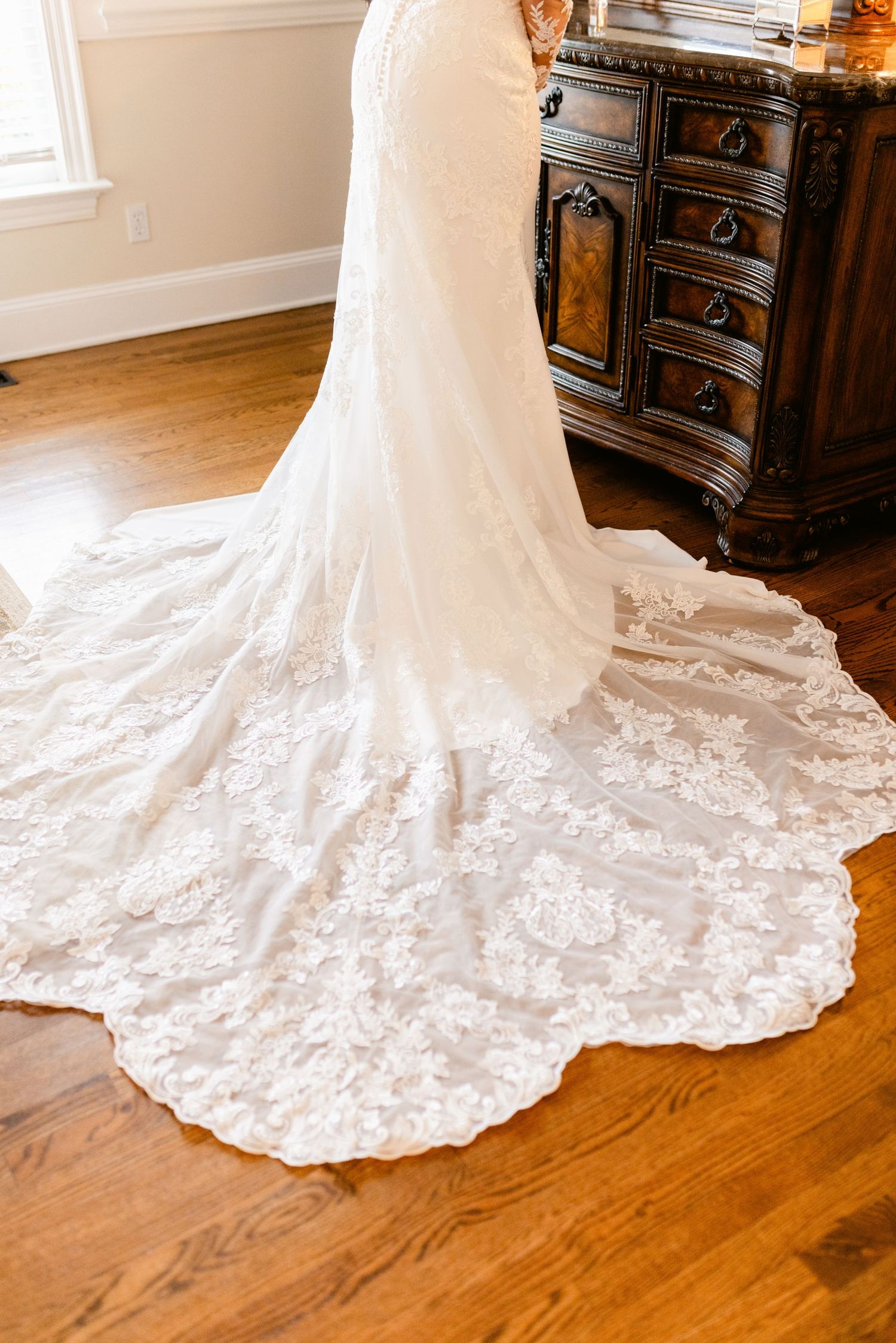 A stunning white lace wedding dress with a long cathedral train displayed on hardwood floors next to an ornate dresser.