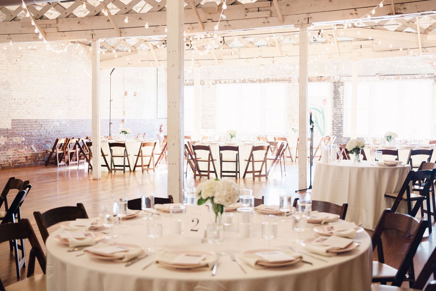 Elegant wedding reception venue with round tables, white linens, and wooden folding chairs in an industrial-style space.