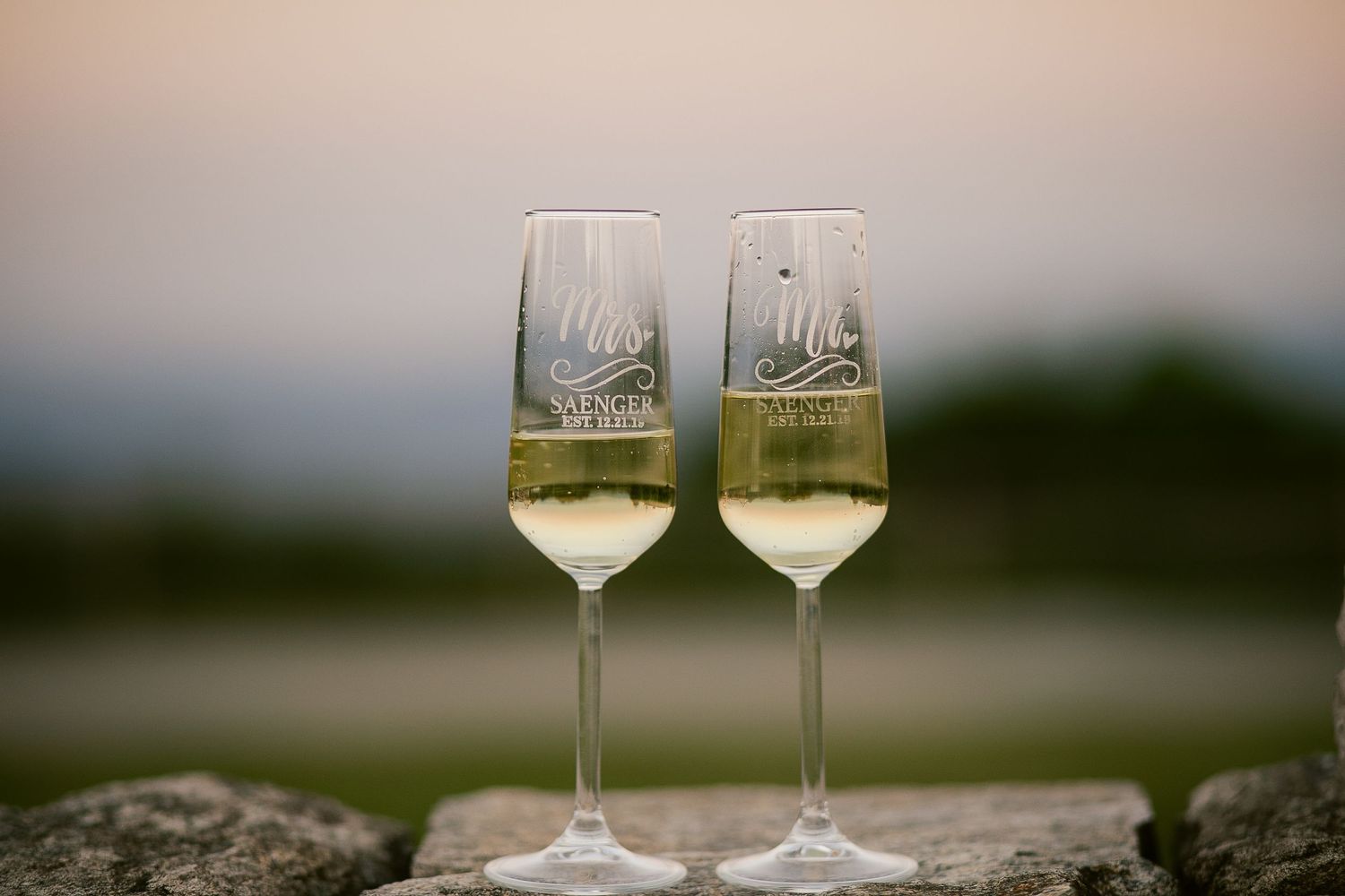 Two champagne flutes with monogrammed designs placed on stone wall at sunset.