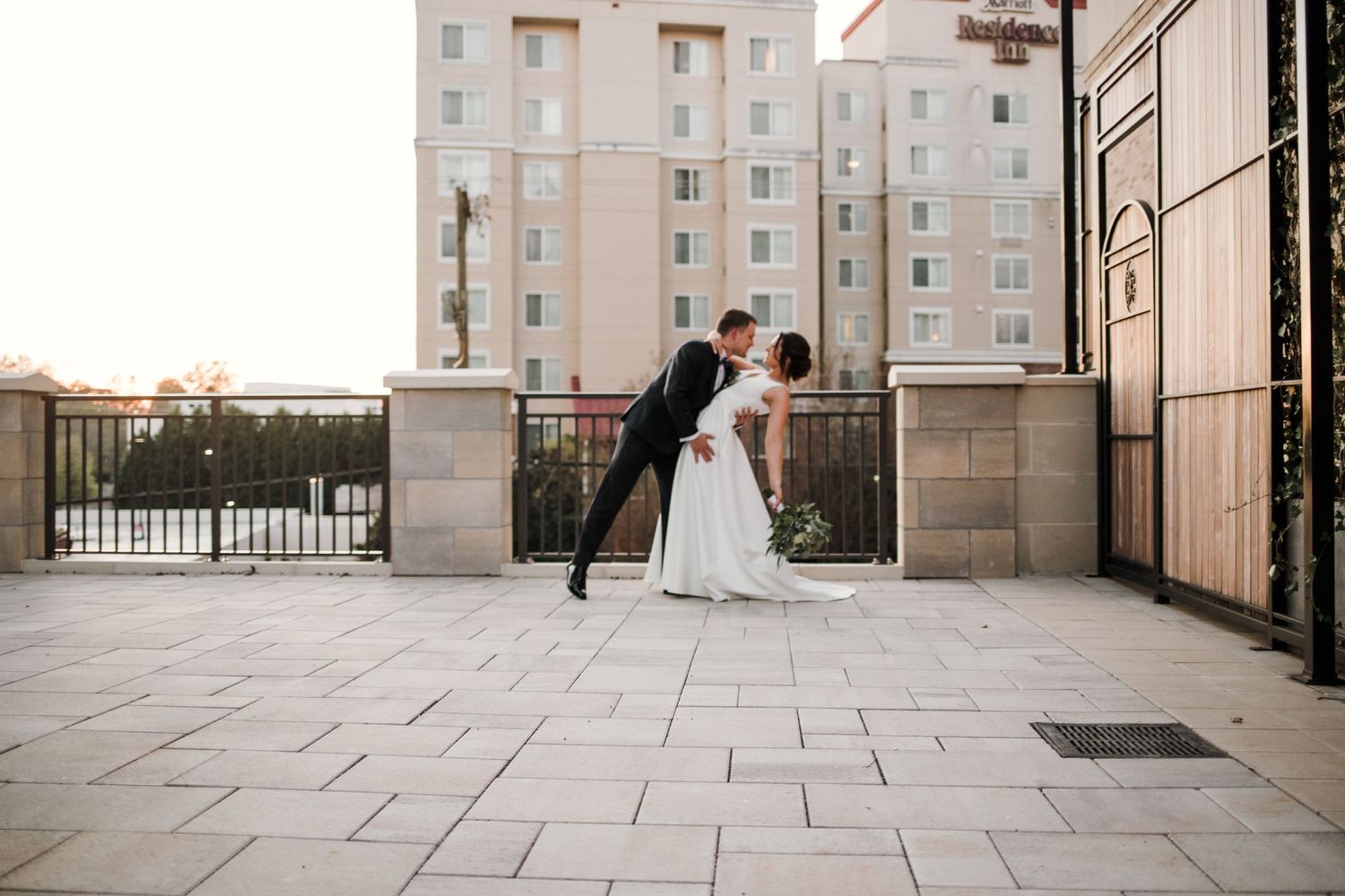 Romantic dip pose between newlyweds on stone patio with city architecture in background.
