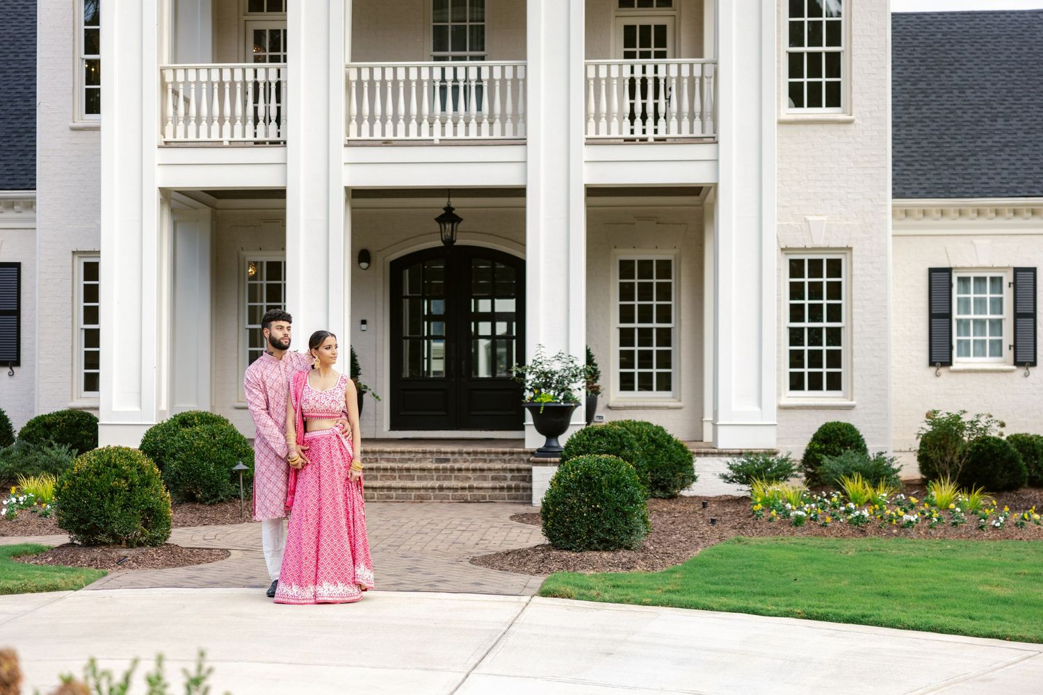 White colonial style mansion with columns and balconies stands behind manicured lawn and landscaping.