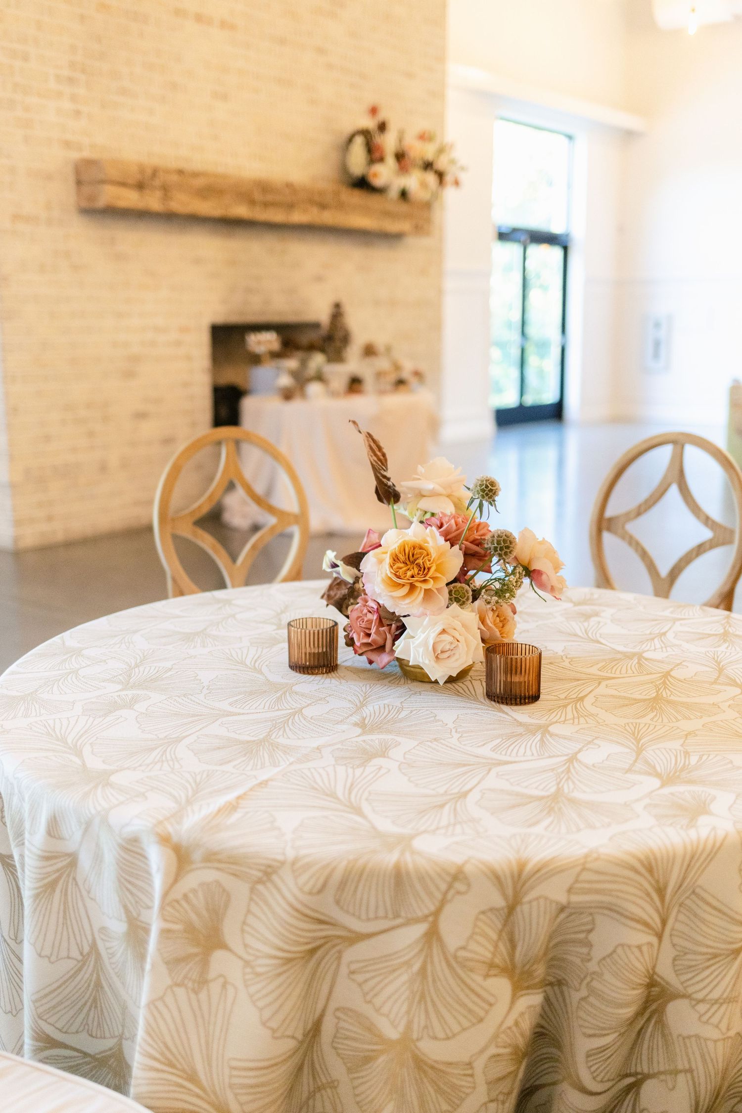 Elegant round reception tables with white textured linens and floral centerpieces in a bright venue with brick walls.
