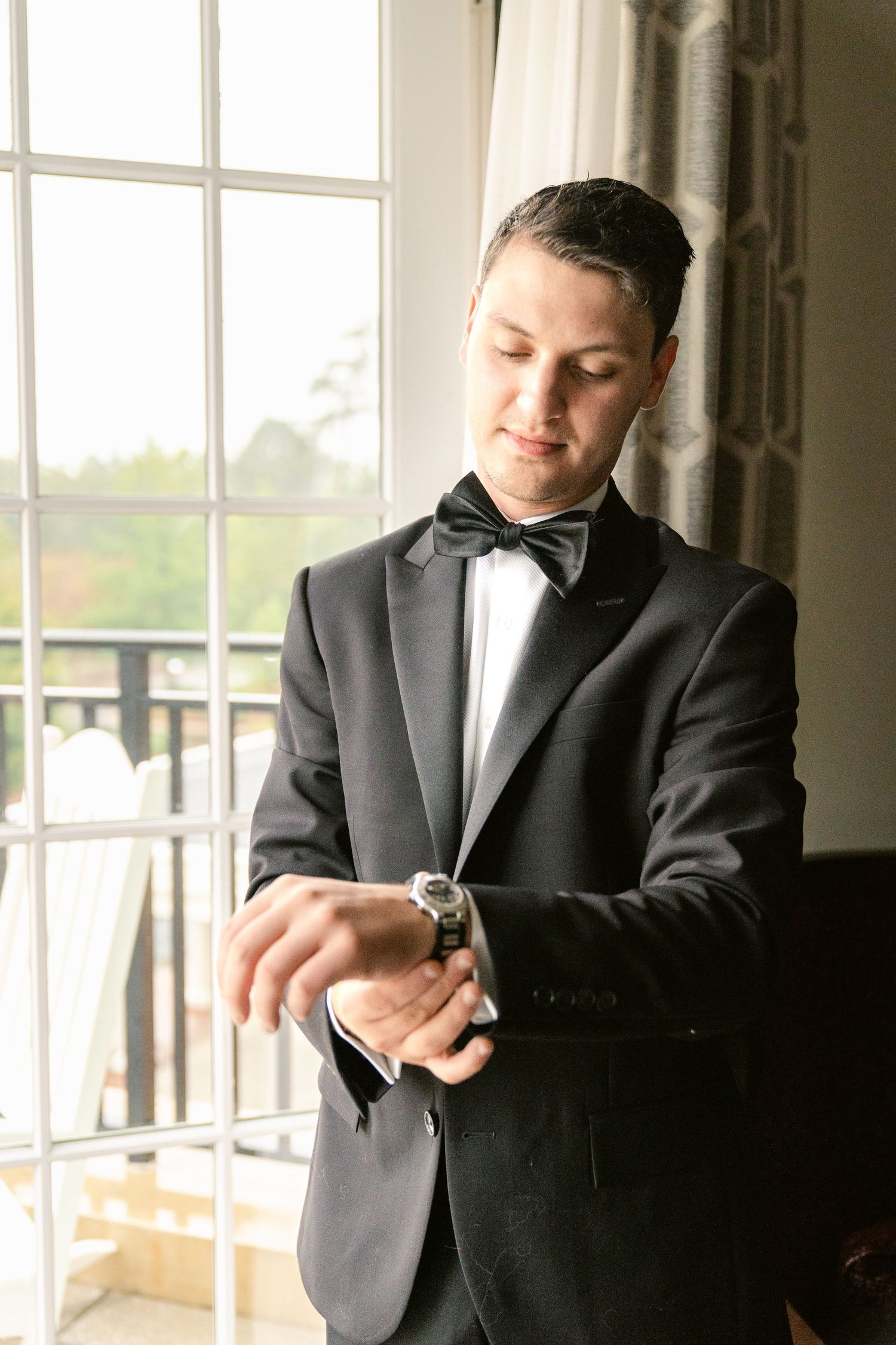 A person in a black tuxedo and bow tie checks their watch while standing by large windows in an elegant room.