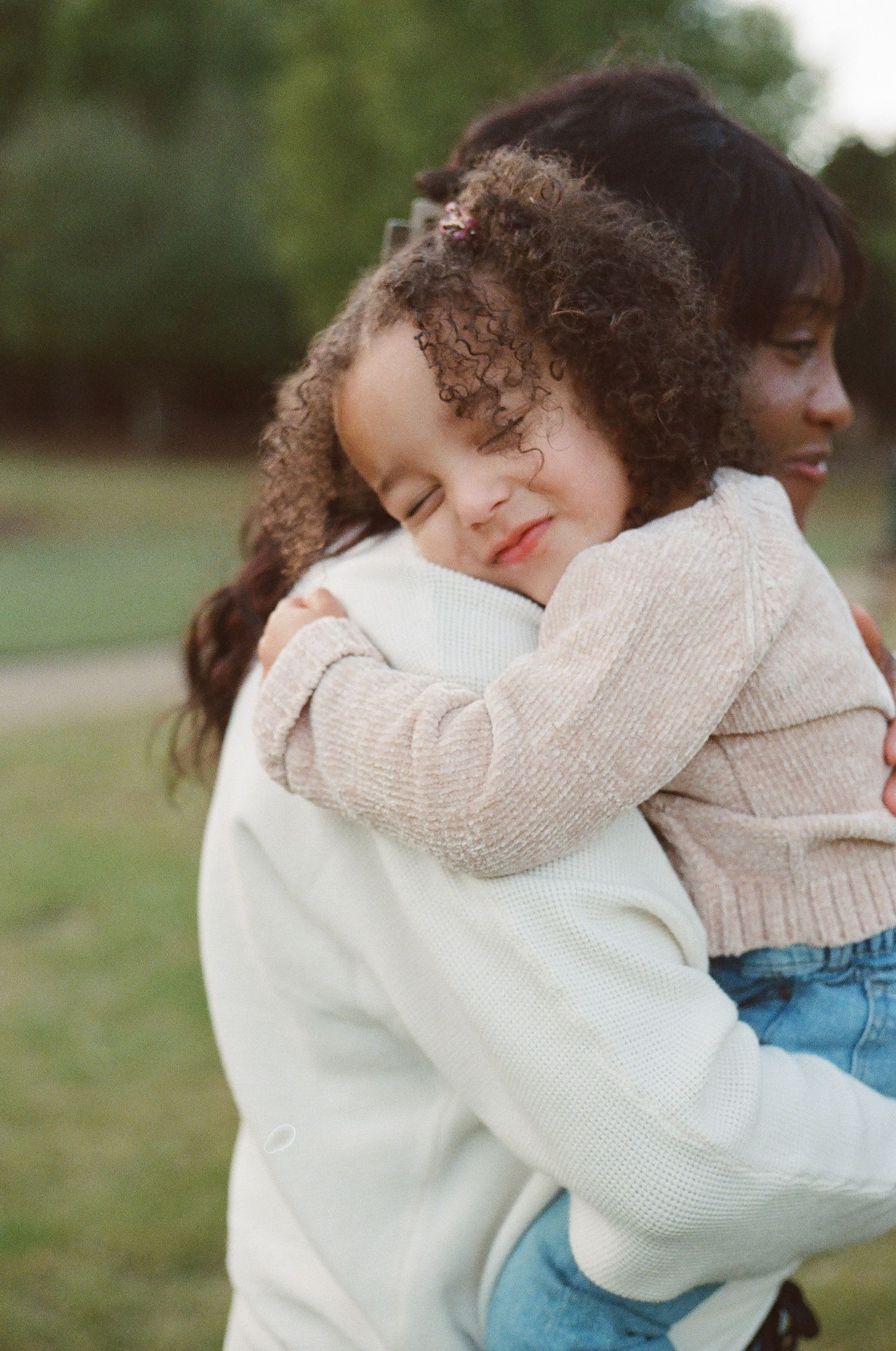 mom and daughter in cozy white and beige sweaters embrace tenderly in an outdoor setting with soft natural lighting on film