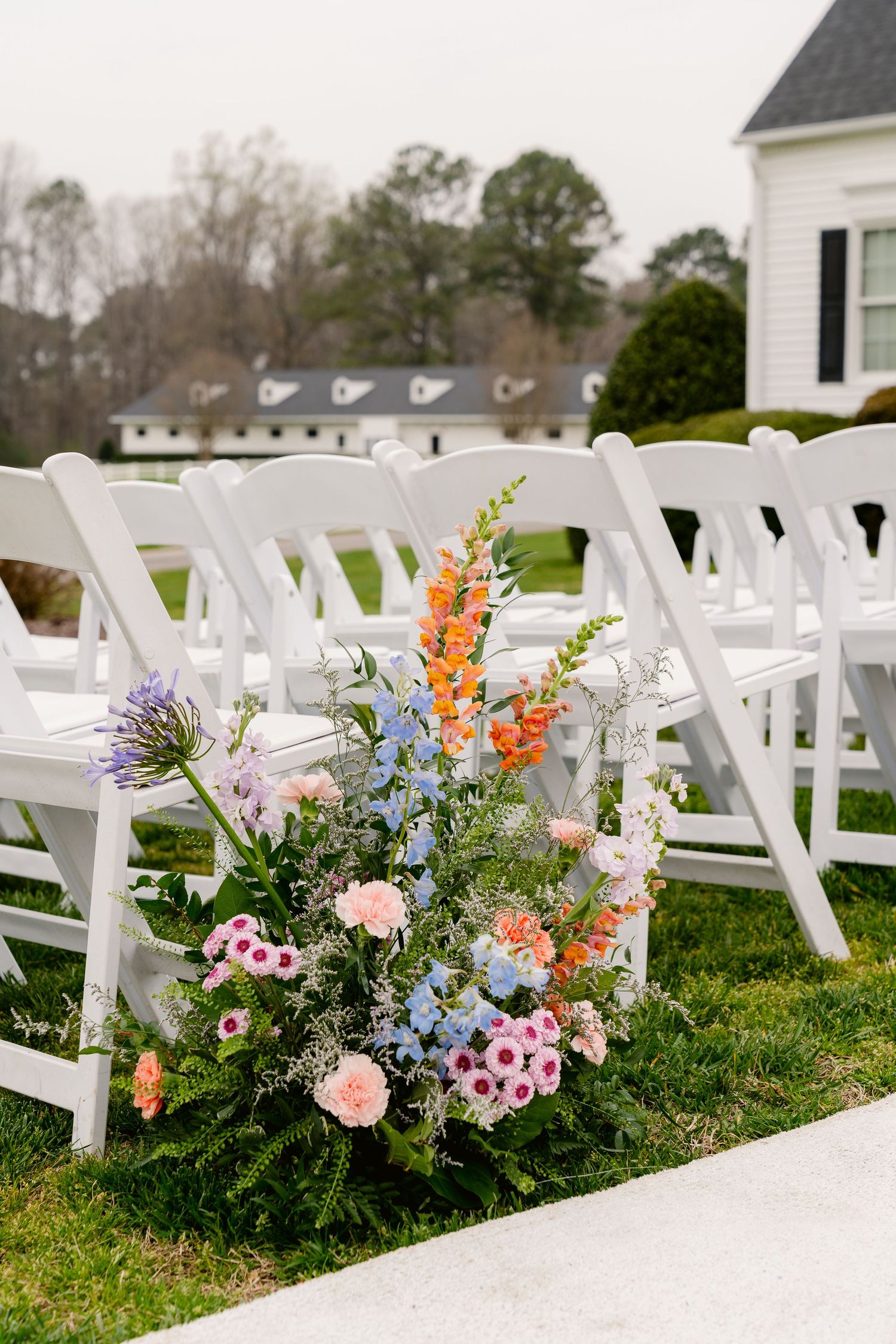 Beautiful outdoor wedding ceremony setup with white chairs and colorful wildflower aisle arrangements in pink, orange and blue.