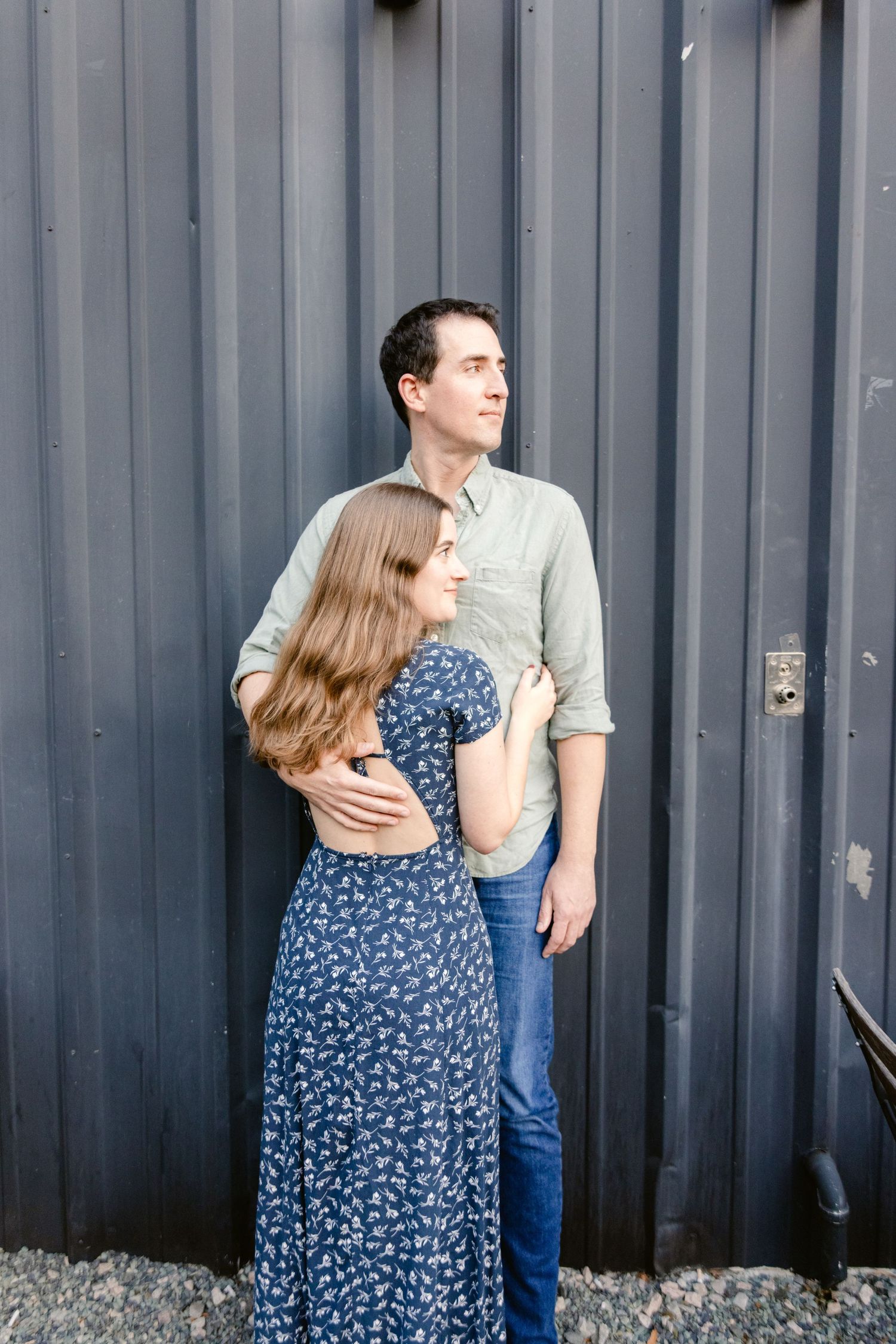 A couple embraces while standing against a grey corrugated metal wall.