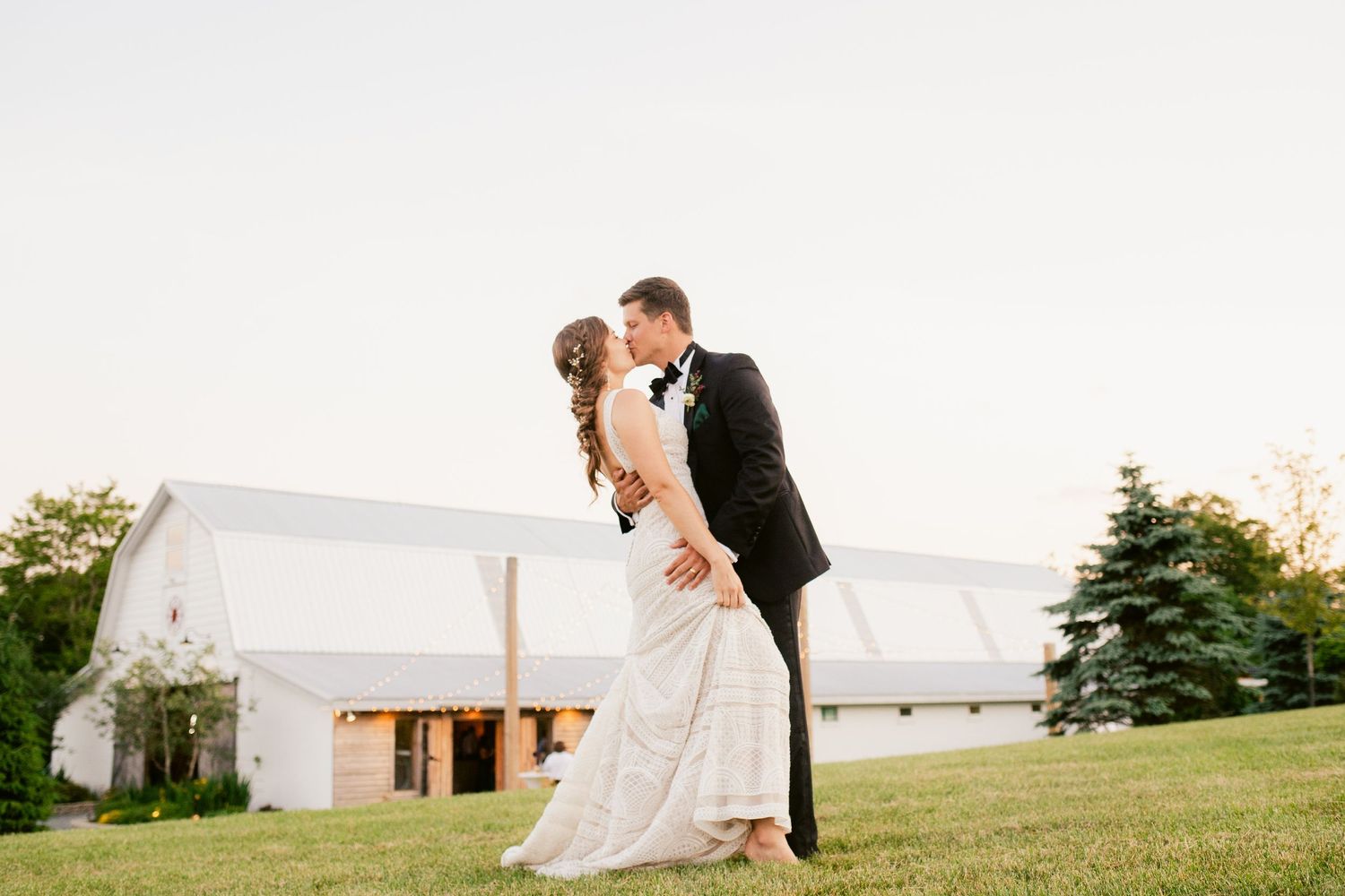 Romantic dip pose by newlyweds on grassy hill with white barn in background.