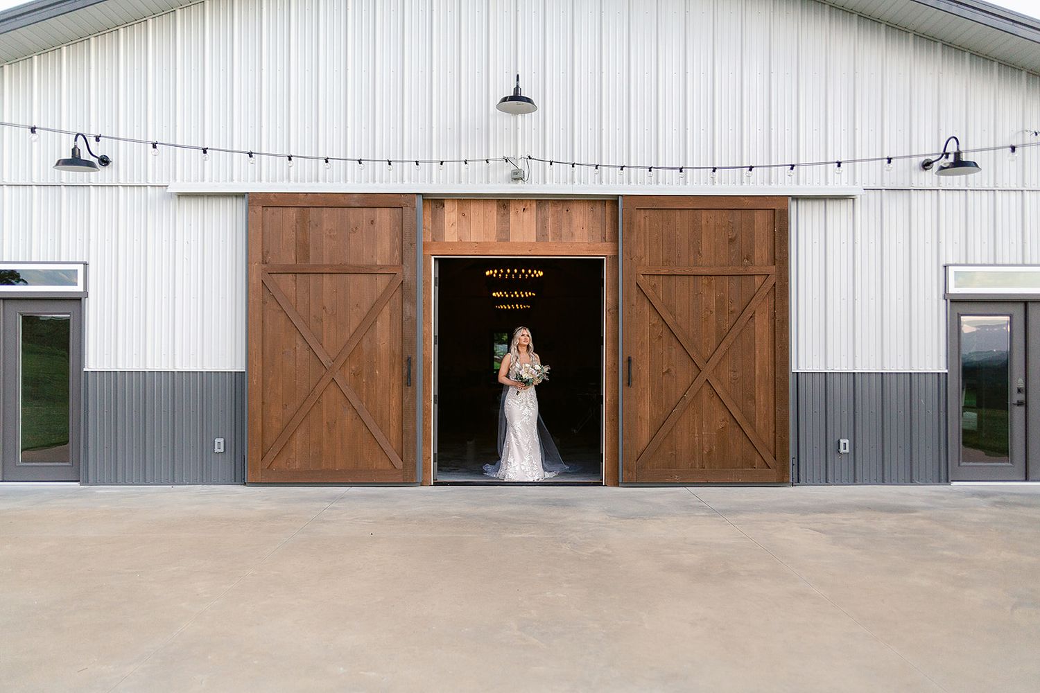 A white barn wedding venue with large wooden sliding doors and a figure in white standing in the doorway.