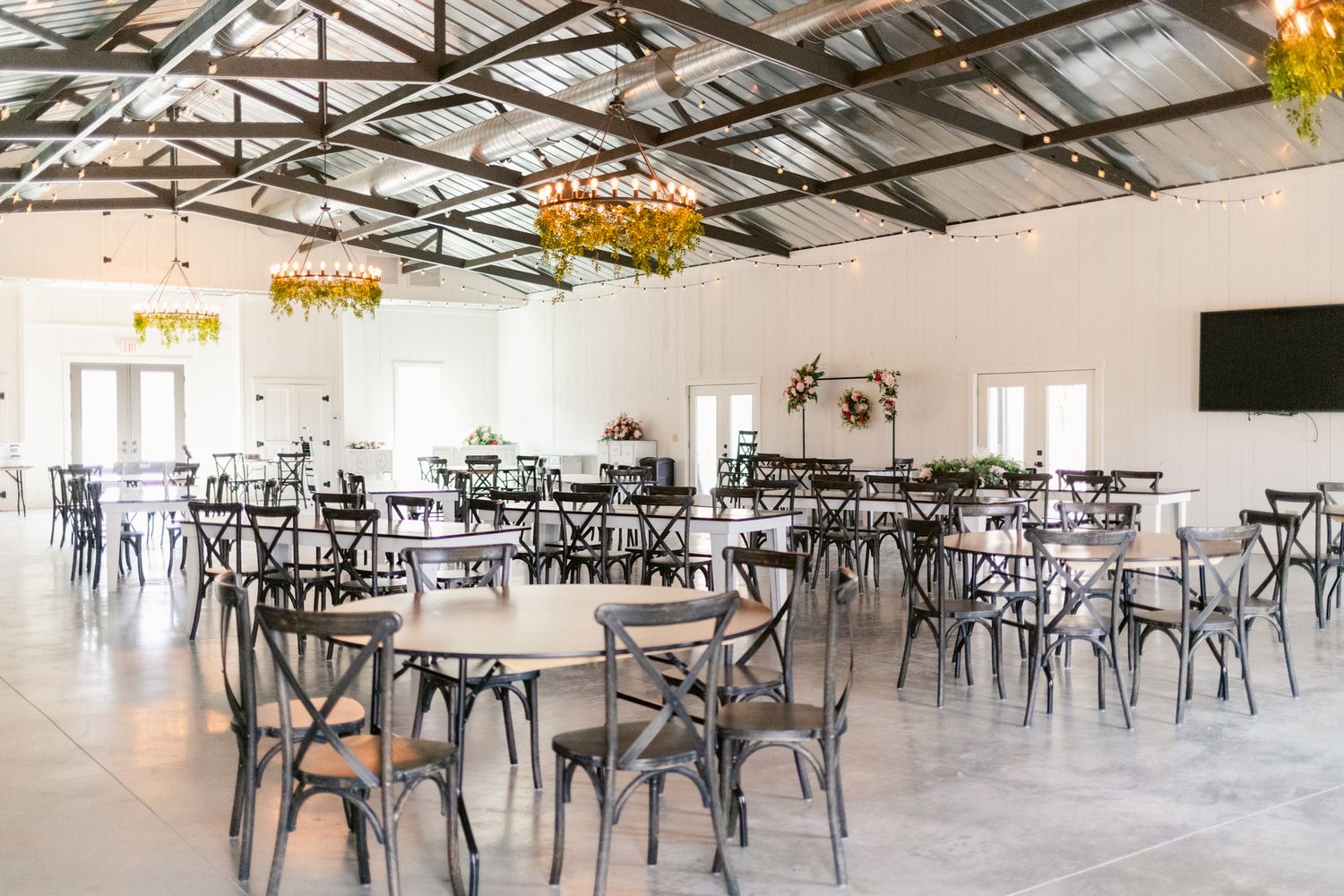 Large rustic reception hall with exposed beams, crystal chandeliers, and black cross-back chairs arranged around round tables.