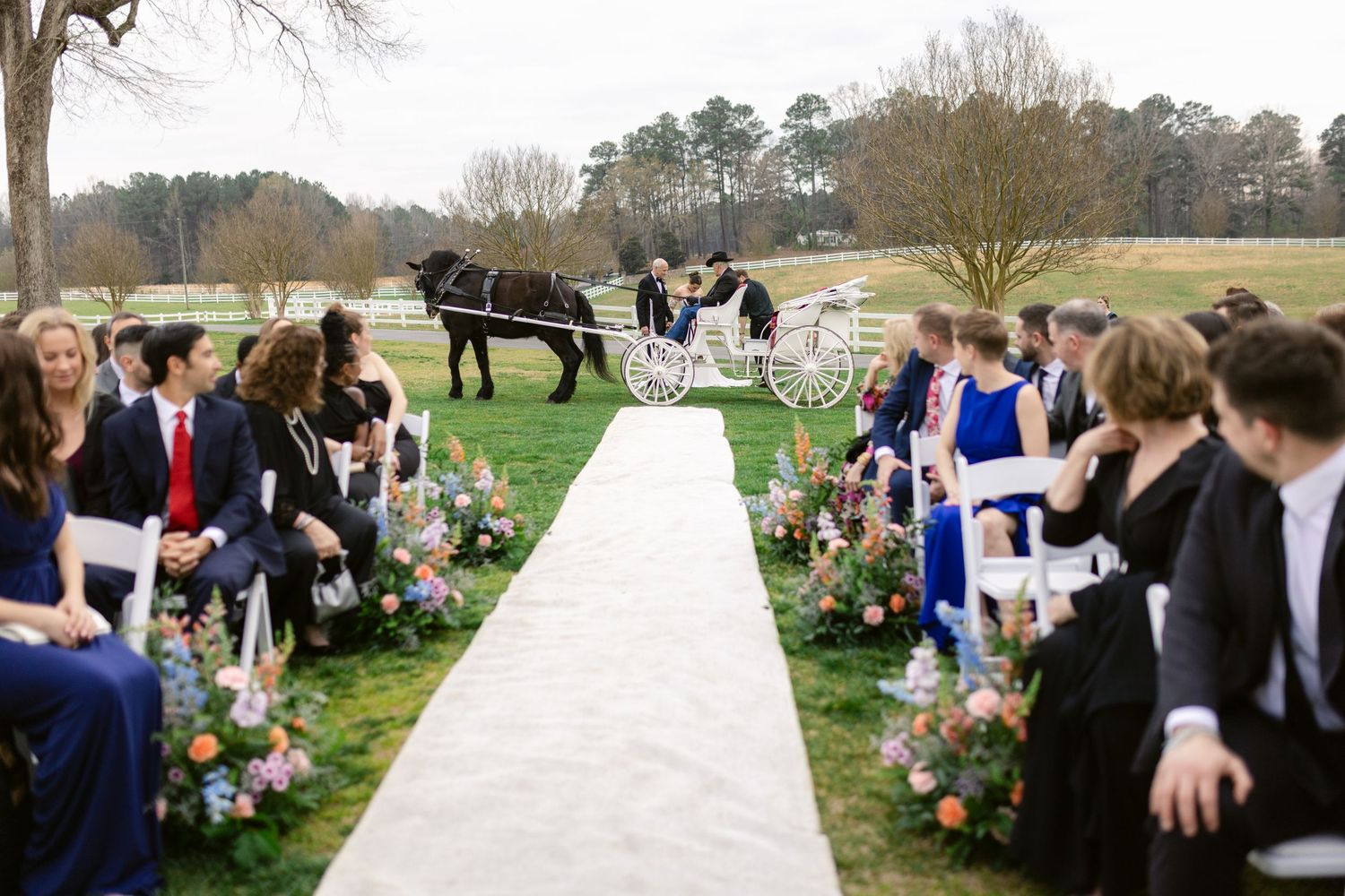 Outdoor wedding ceremony setup with white aisle runner, floral decorations, and horse-drawn carriage in countryside setting.