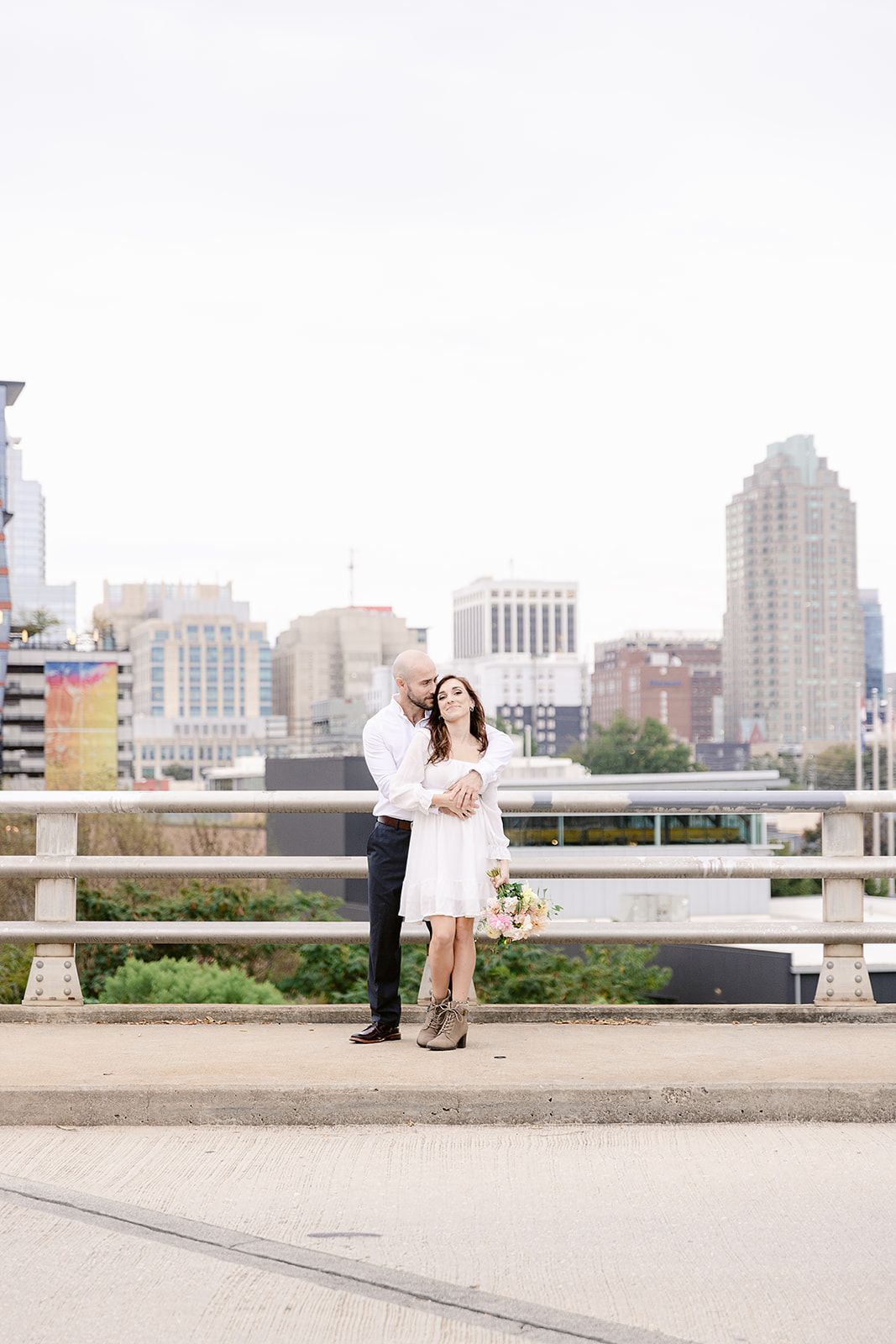 A couple shares a romantic moment on a rooftop terrace with an urban cityscape backdrop.