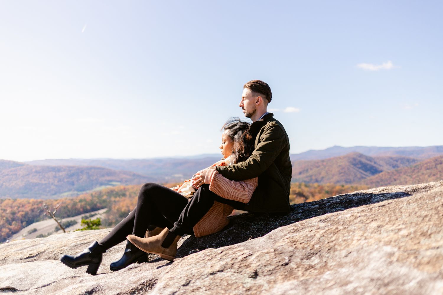 A person sits peacefully on a rocky mountain ledge overlooking a vast autumn landscape.
