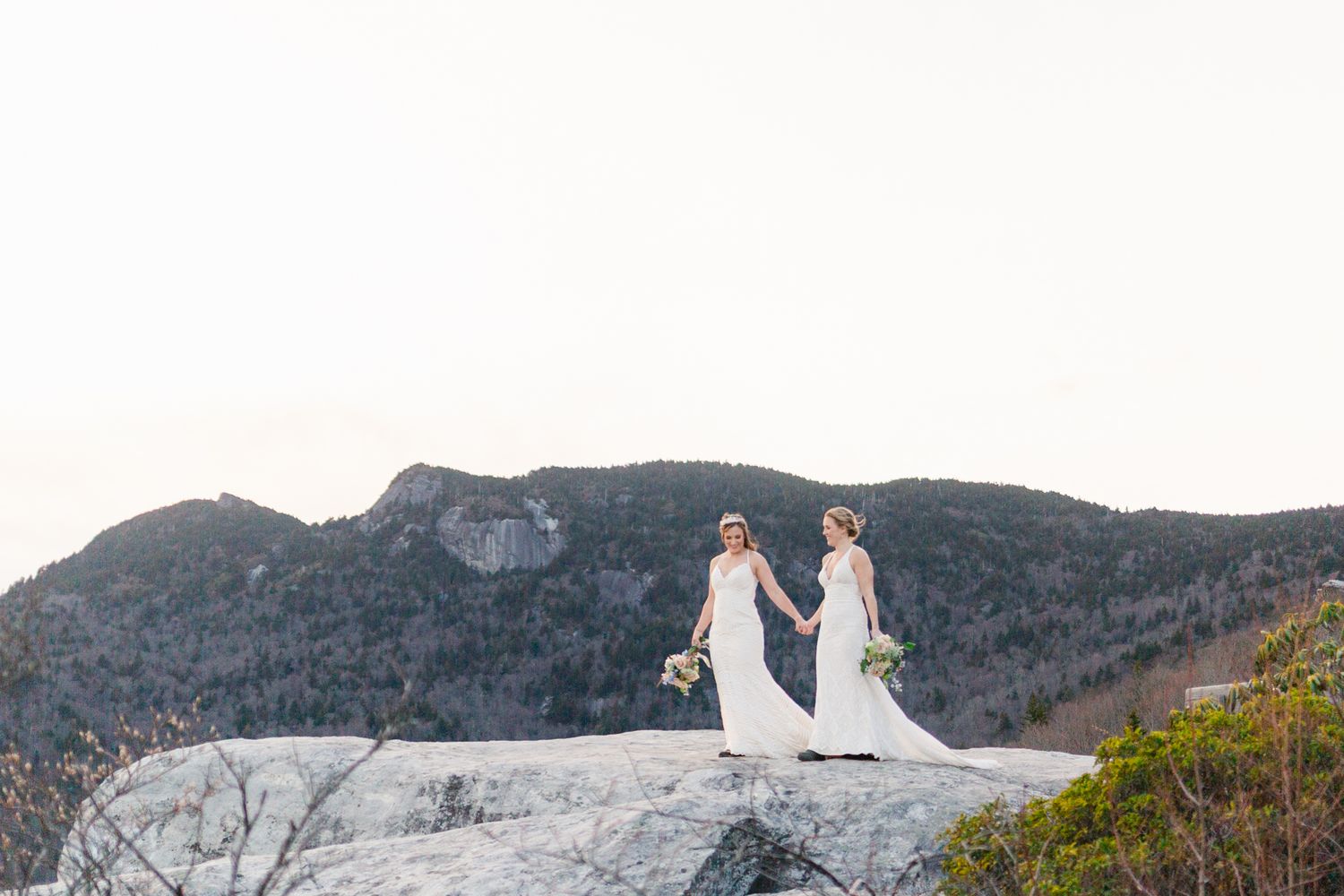Two people in white dresses standing on a rocky mountain overlook.