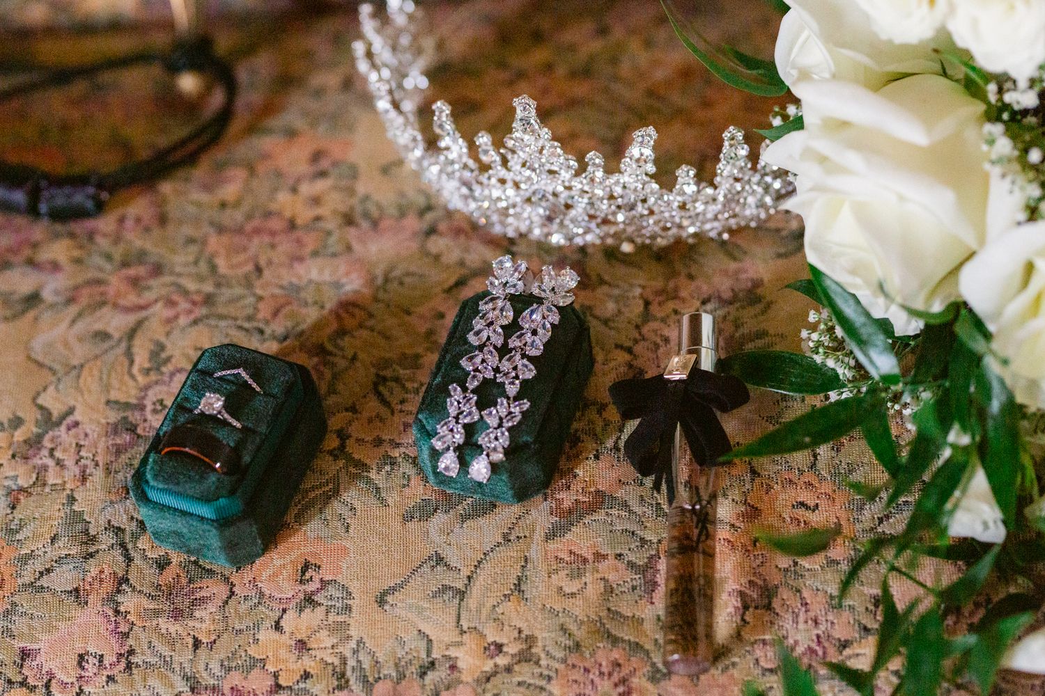 Wedding accessories including a crystal tiara, white roses, and delicate jewelry arranged on a pink floral surface.
