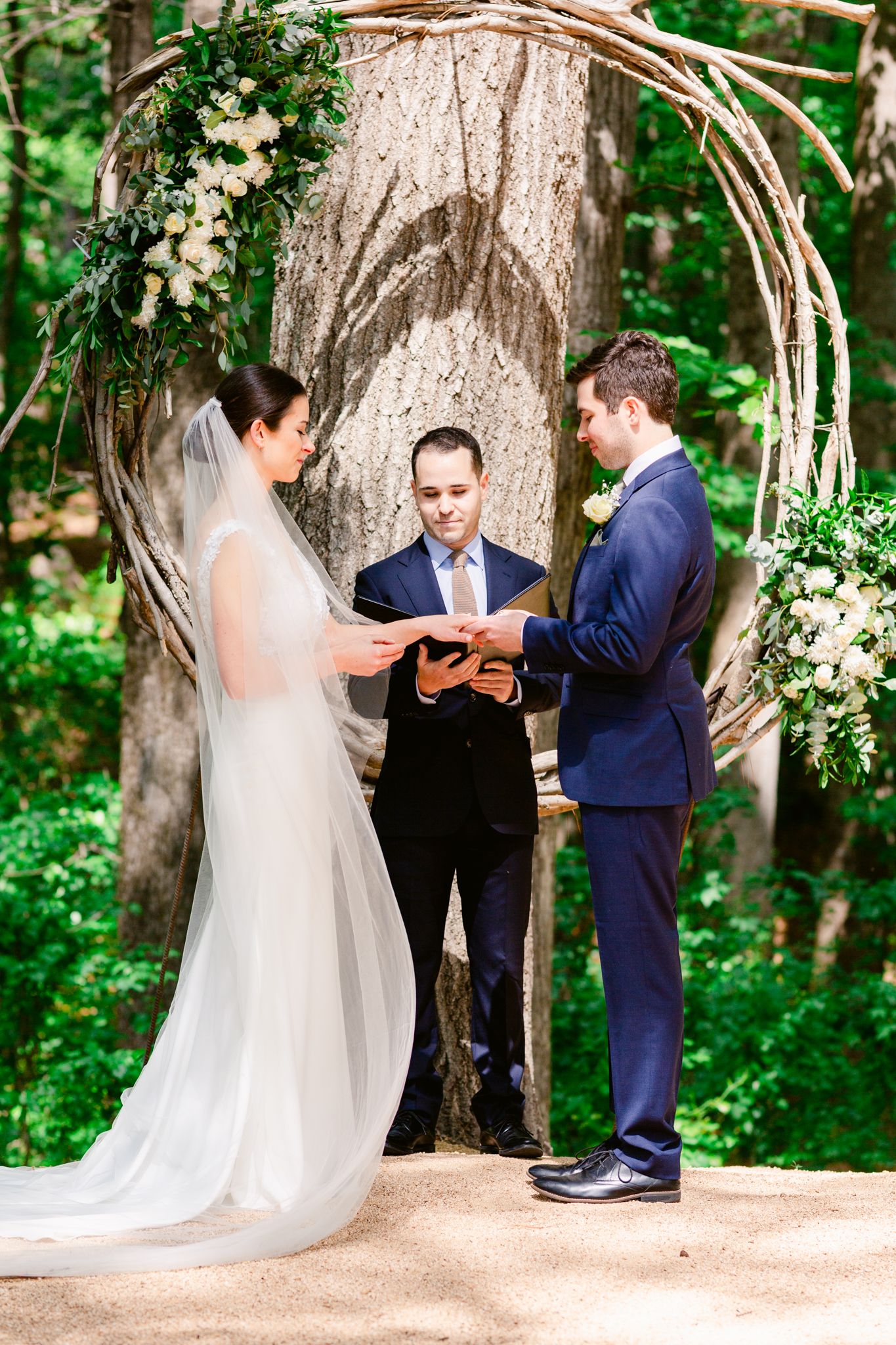 A romantic outdoor wedding ceremony takes place under a rustic wooden arch decorated with white floral arrangements.